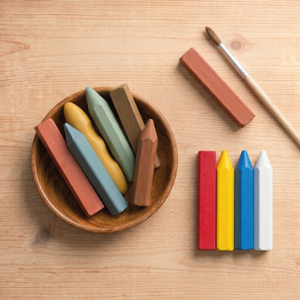 Verschillende gekleurde pastel krijtjes op een houten tafel, met een houten kom en een verfkwast naast.