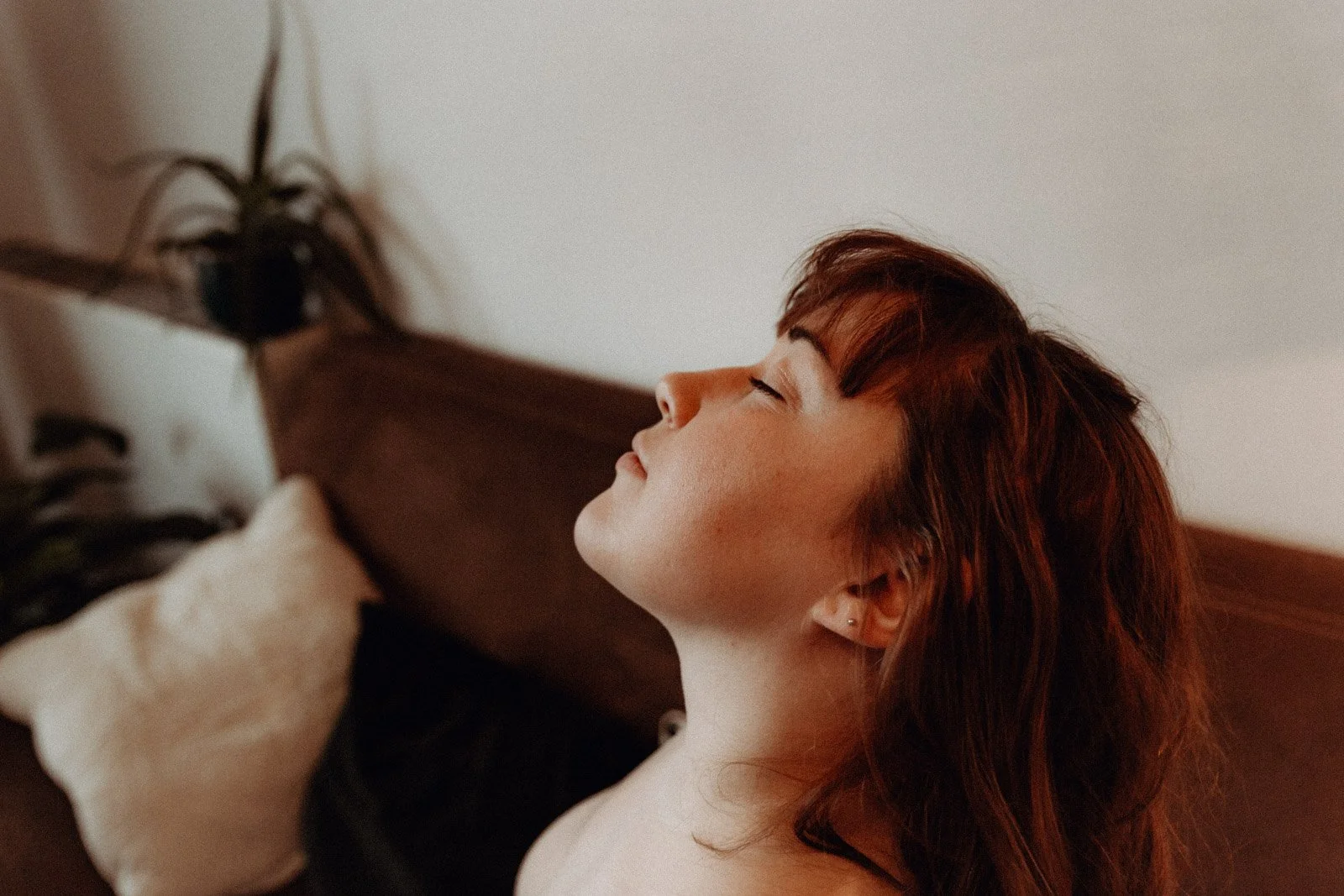 Femme aux cheveux bruns, yeux fermés, tête inclinée en arrière, plante et coussin en arrière-plan.