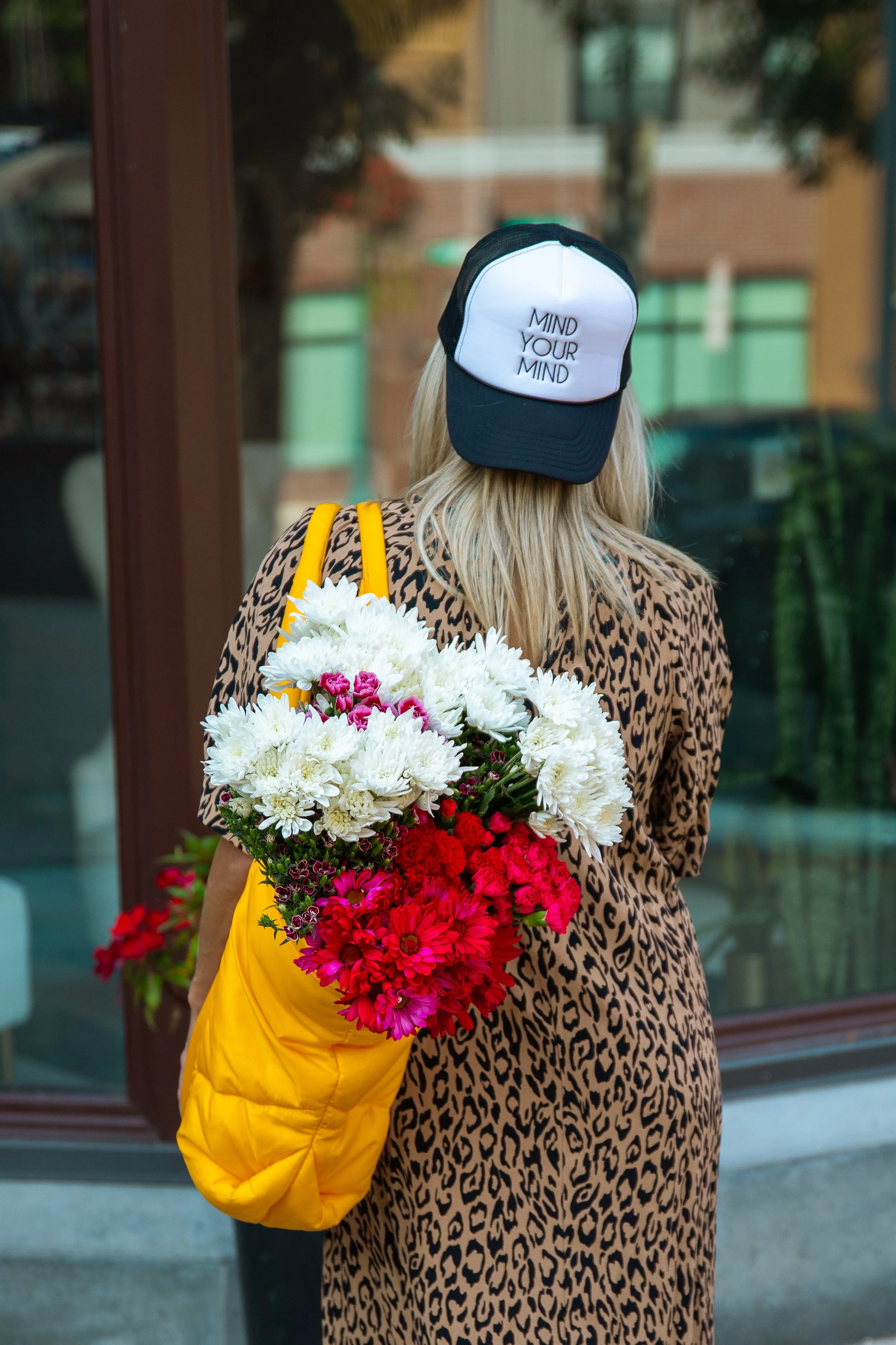 A woman wearing a leopard print dress, a black and white cap with text, and carrying a yellow bag filled with white and pink flowers, standing outside near a glass window.