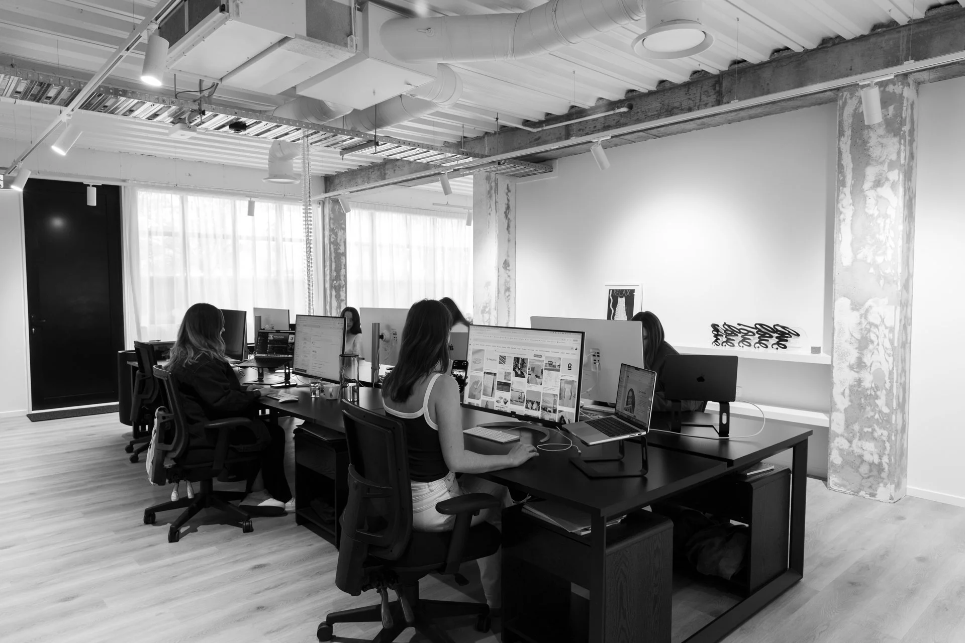George Design open-plan office with four women working at desks with computers, set in a modern space with large windows, sheer curtains, exposed ceiling pipes, and minimalist decor in black and white.