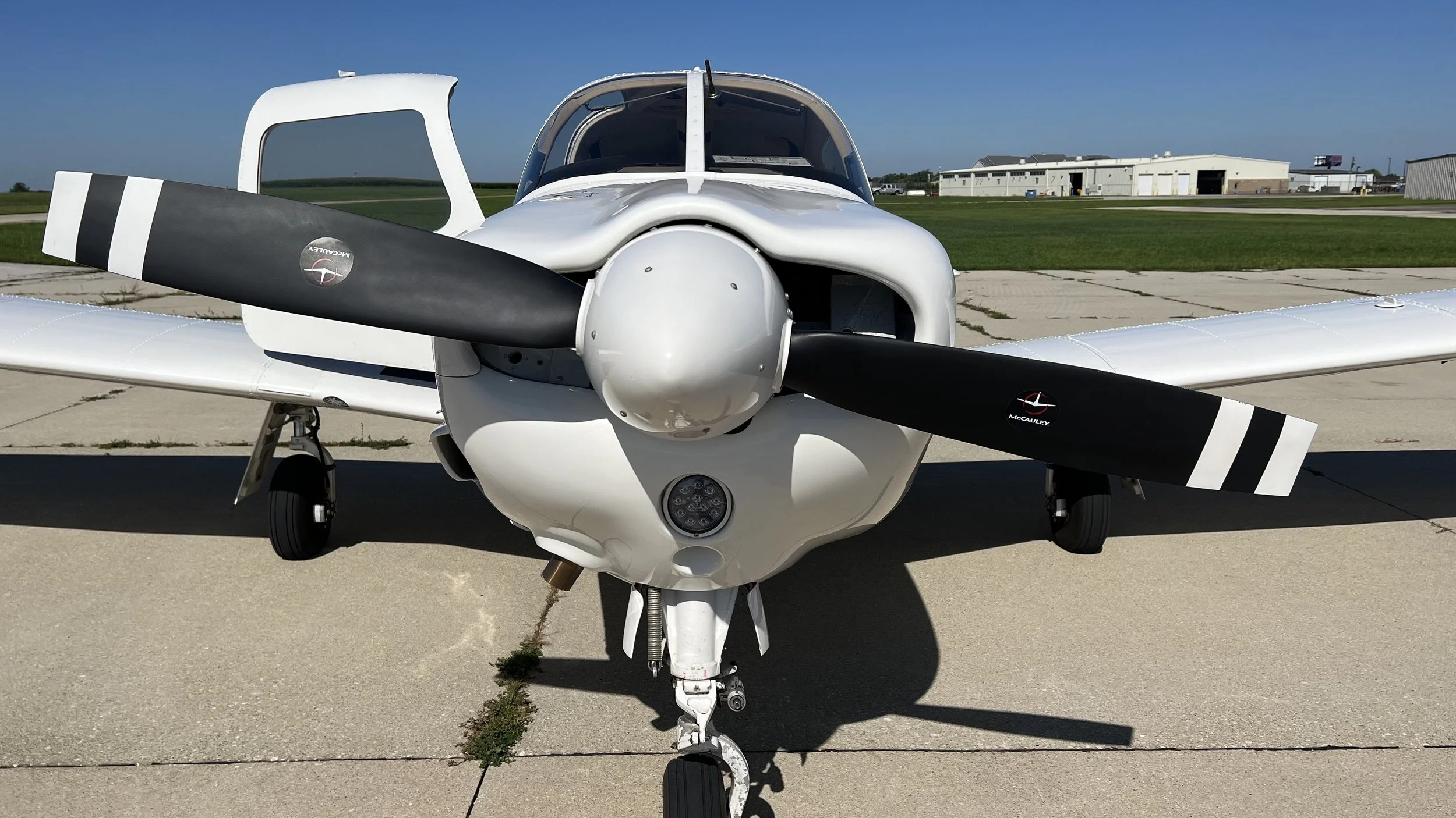 Front view of a white single-engine airplane with a black propeller on an airport tarmac under a clear blue sky.