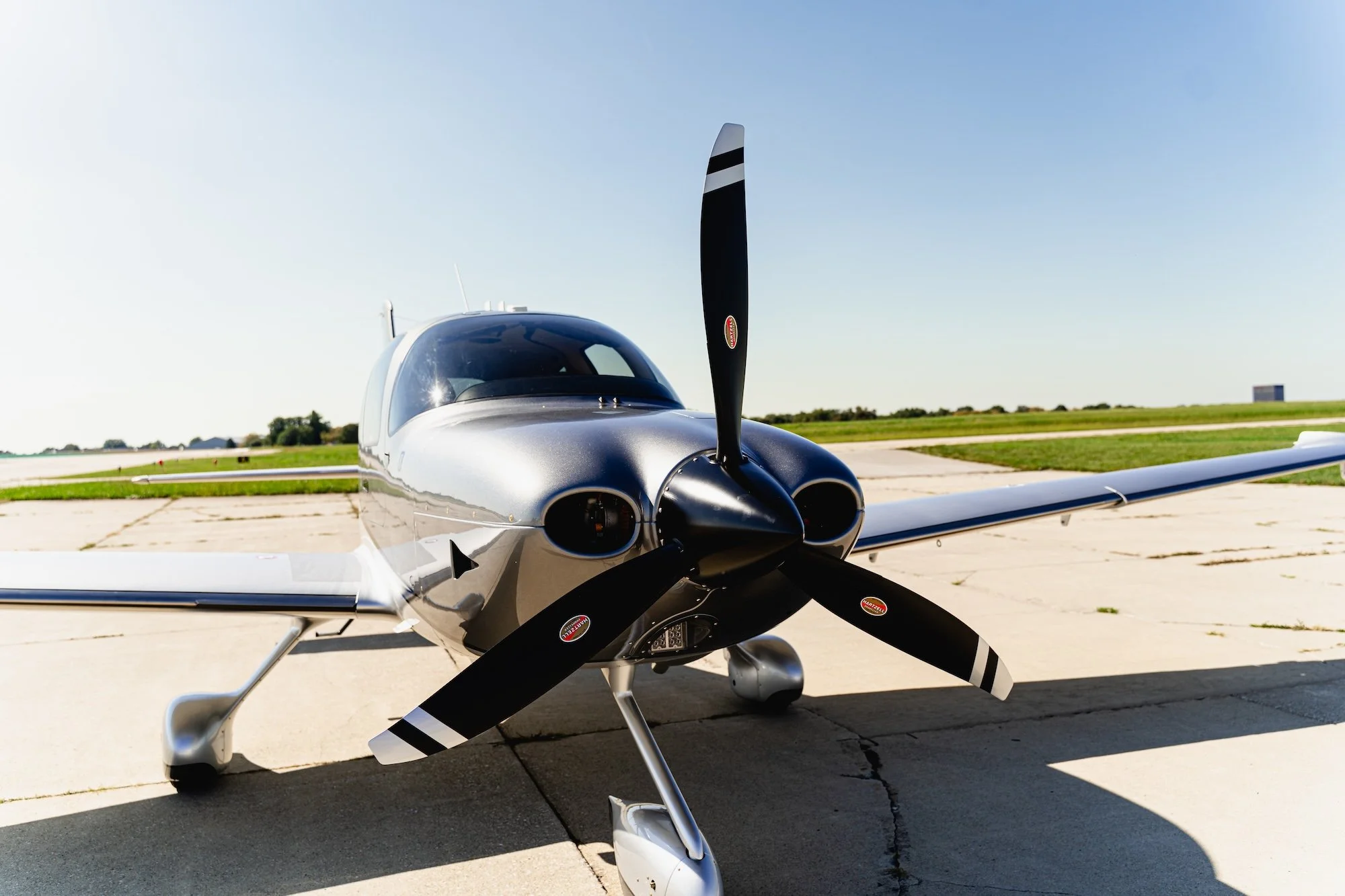 A small, silver and black airplane parked on a concrete runway on a sunny day with a green grass field in the background.