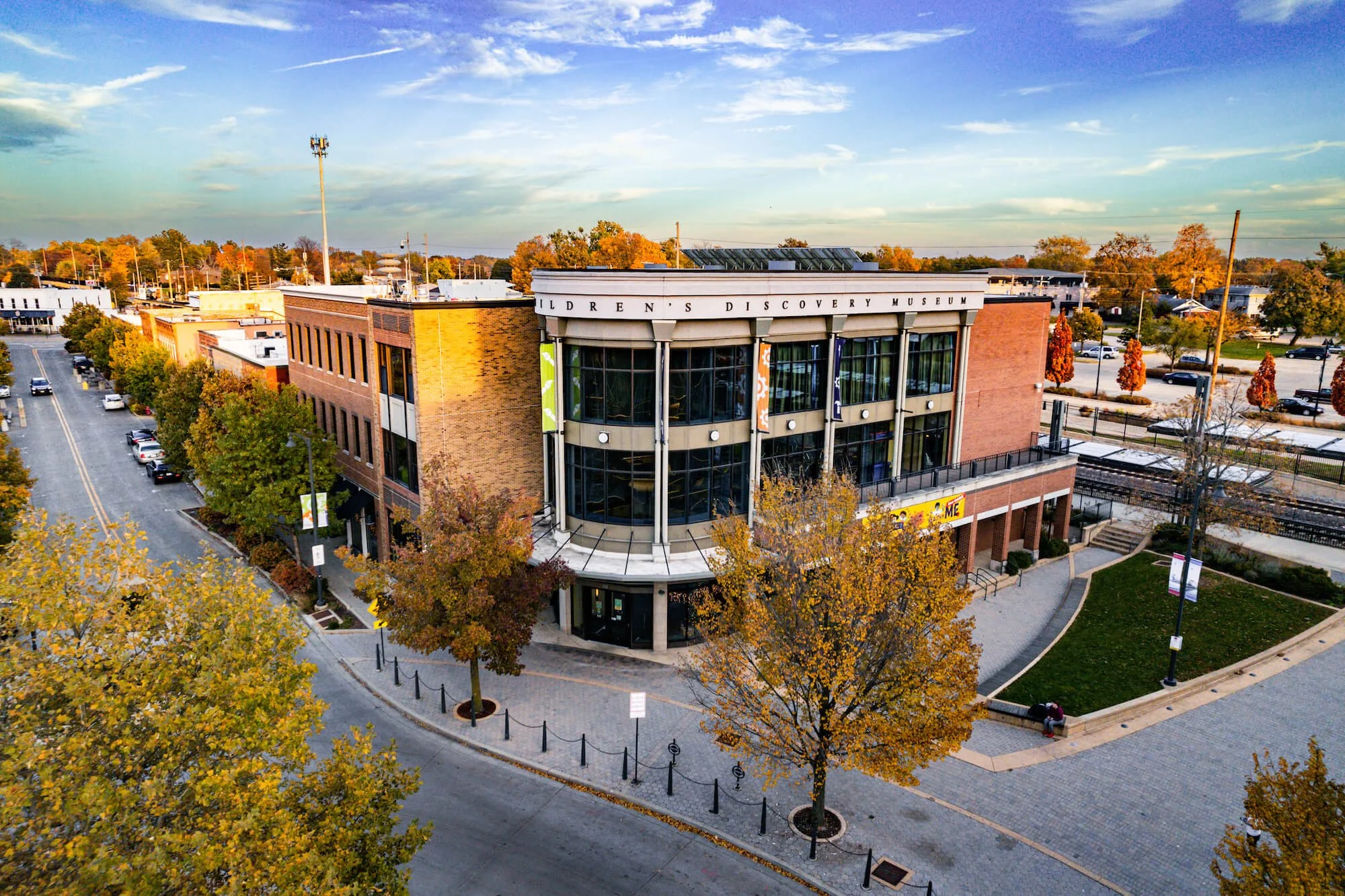 An aerial view of a city block with a modern building labeled 'Children's Discovery Museum,' surrounded by trees with autumn foliage, parking lots, and streets under a partly cloudy sky.