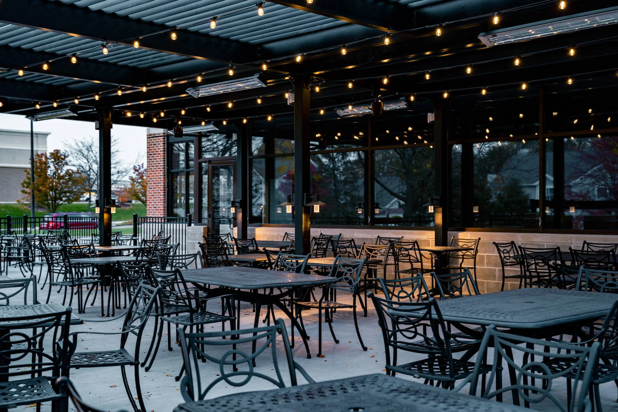 Empty outdoor patio with black metal tables and chairs, string lights hanging overhead, enclosed by glass walls, during dusk or early evening.