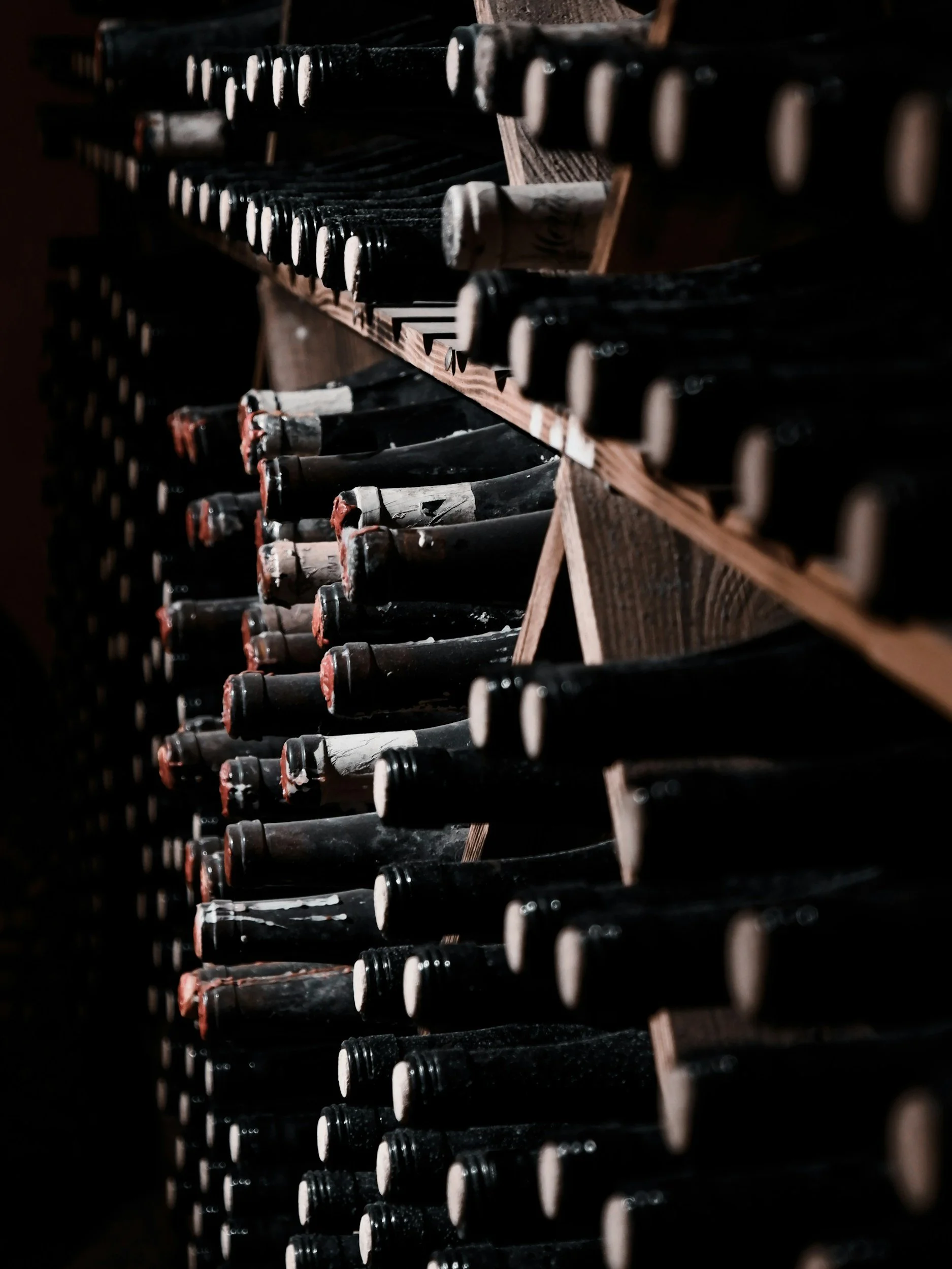 Una fila di bottiglie di vino disposte orizzontalmente in una cantina buia, con alcune etichette e colli visibili.