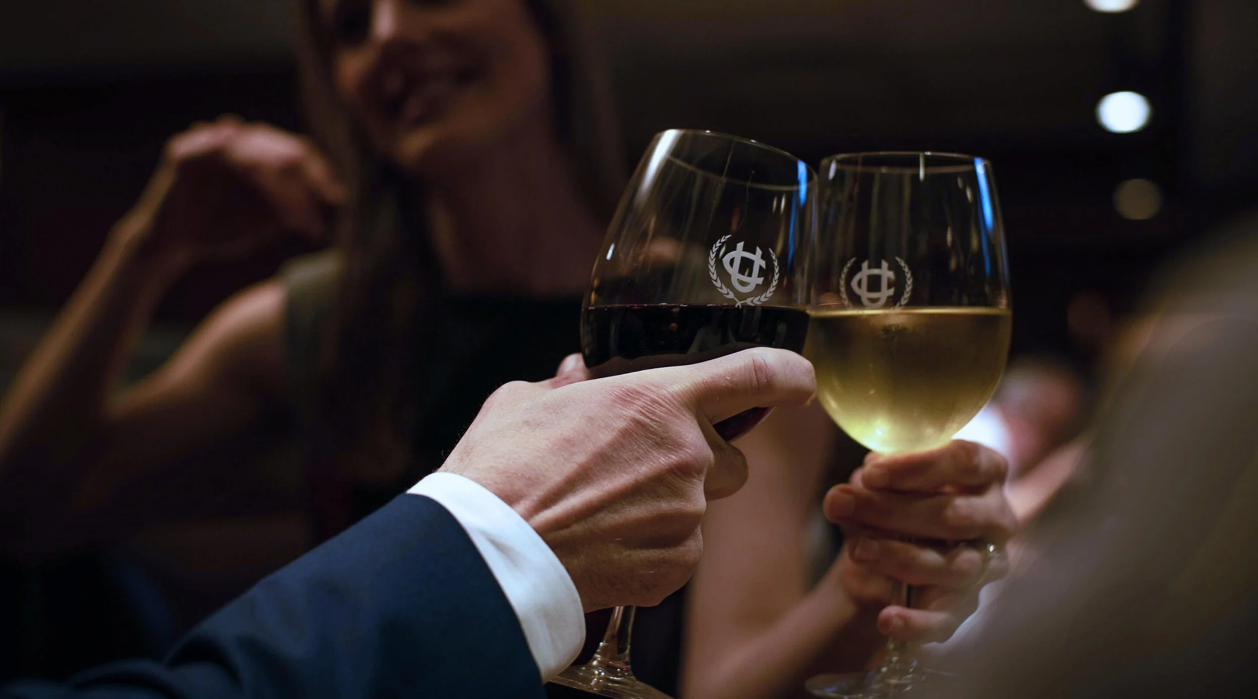 Close-up of two people clinking glasses of red and white wine at a gathering or celebration, with a woman smiling in the background.