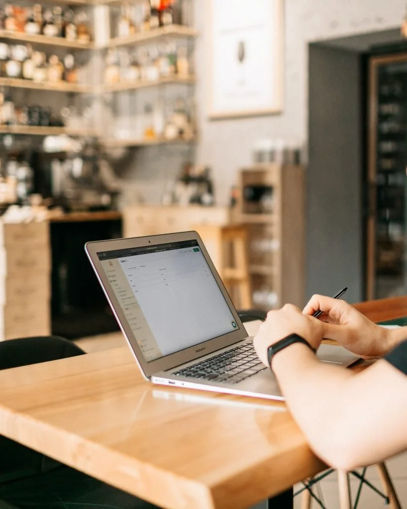 Una persona seduta a un tavolo di legno davanti a un MacBook argento con un grafico sullo schermo, in un accogliente caffè con scaffali in legno e uno sfondo sfocato.
