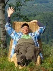 A smiling child sitting outdoors in a wicker chair with arms raised in a peaceful, green landscape.