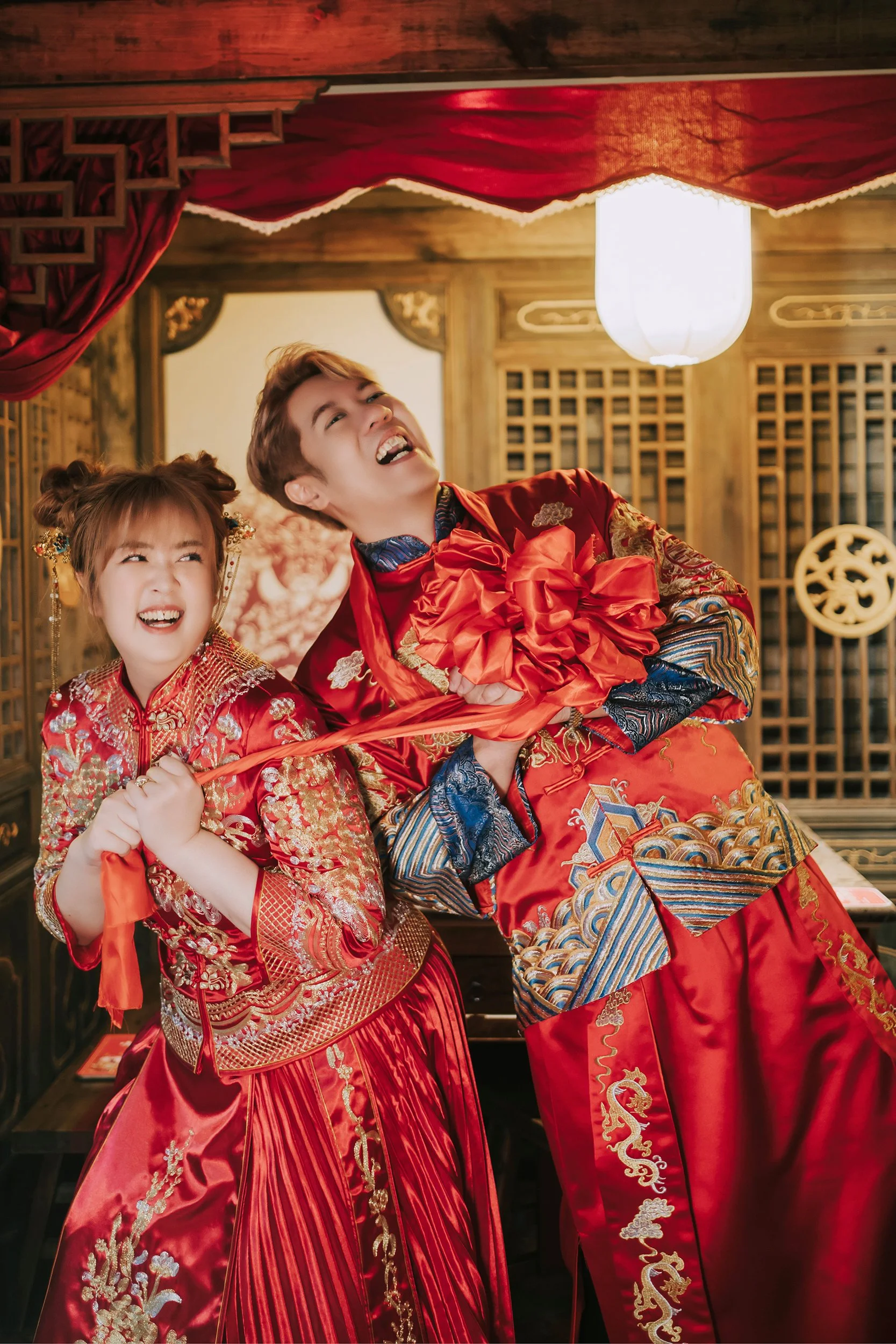 In the atmospheric confines of a traditional Chinese restaurant, a snapshot captures the essence of love and mirth, thanks to the adept lens of Chris Ling Photography. The couple, adorned in striking red Chinese kua, are the epitome of cultural elega