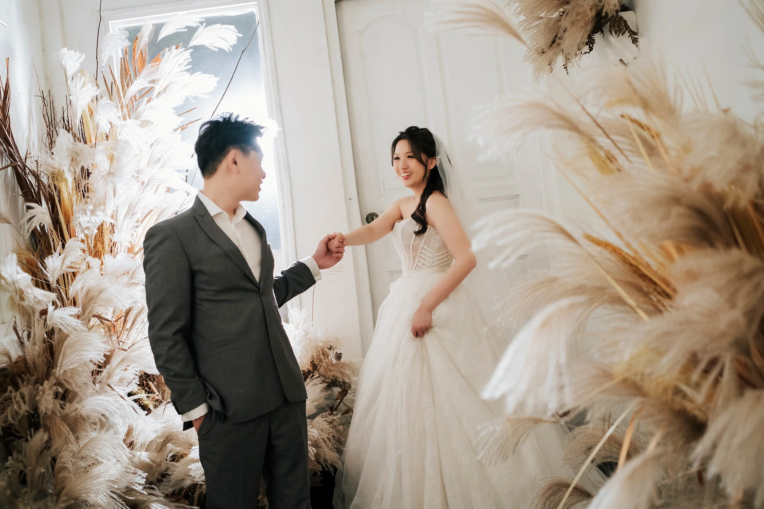 In the sophisticated setting of Chris Ling Photography Studio, a couple in full wedding attire are captured in a moment of soft, timeless romance. Contrasted against a clean white background, the scene is warmly accented with artistically arranged dr