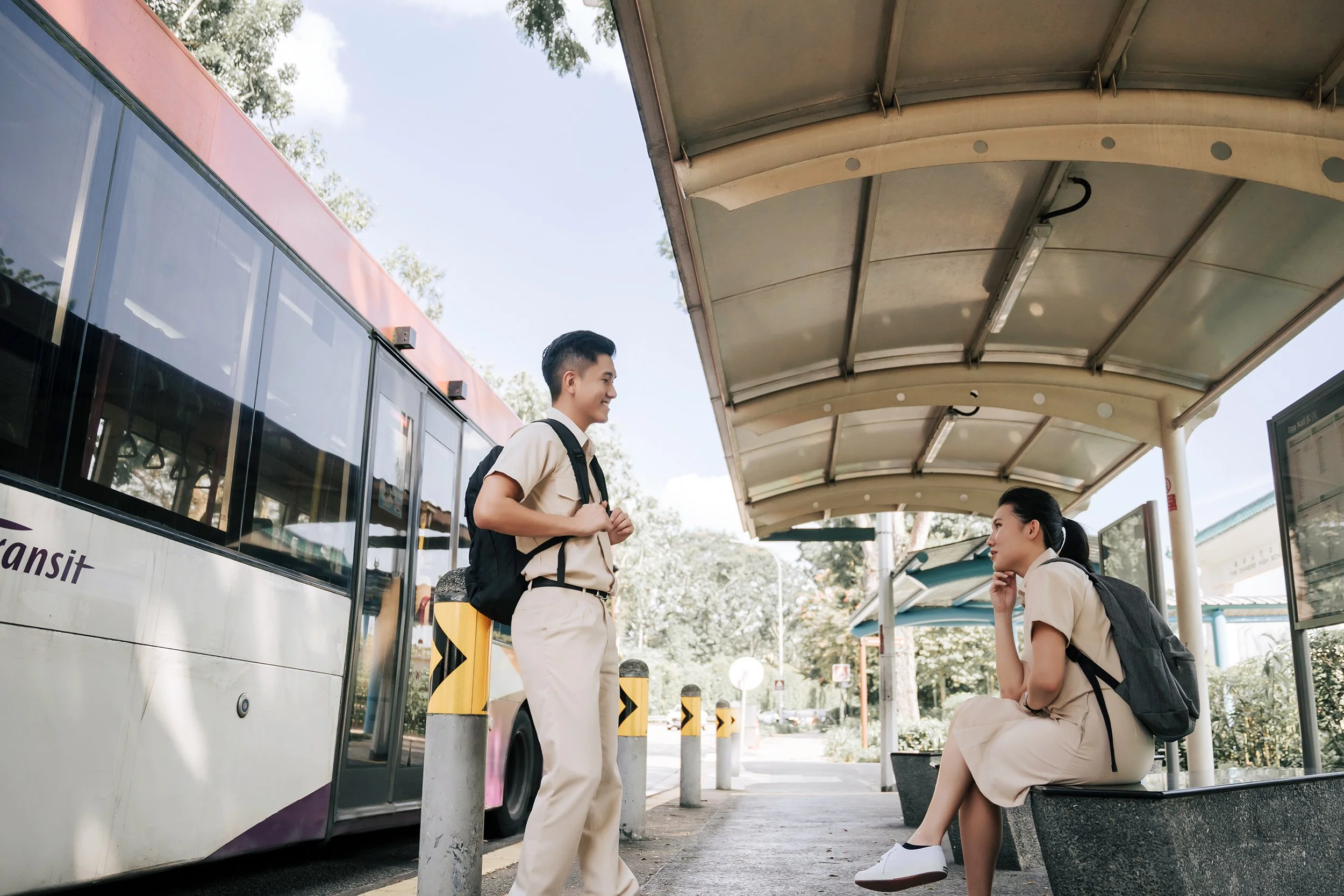 A nostalgic pre-wedding photograph of a couple dressed in matching school uniforms at a bus stop, recreating the innocence and simplicity of young love. With a bus parked beside them and sunlight streaming in, this charming scene captures the beginni