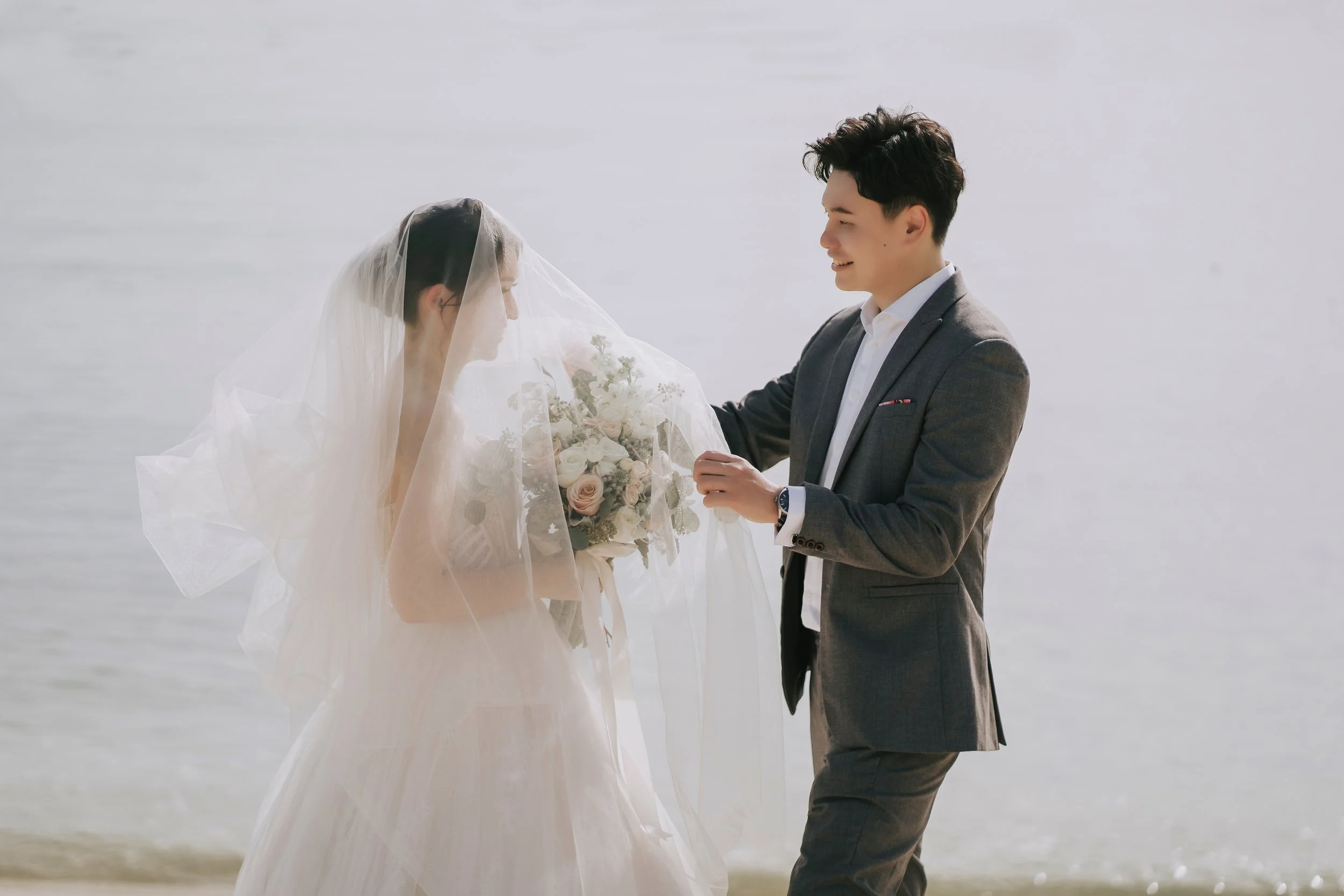 In an idyllic beach setting, a couple celebrates their upcoming nuptials with a captivating pre-wedding photoshoot, juxtaposing the casual setting of the sea with the formality of their full wedding attire. The groom, impeccably dressed in a tailored