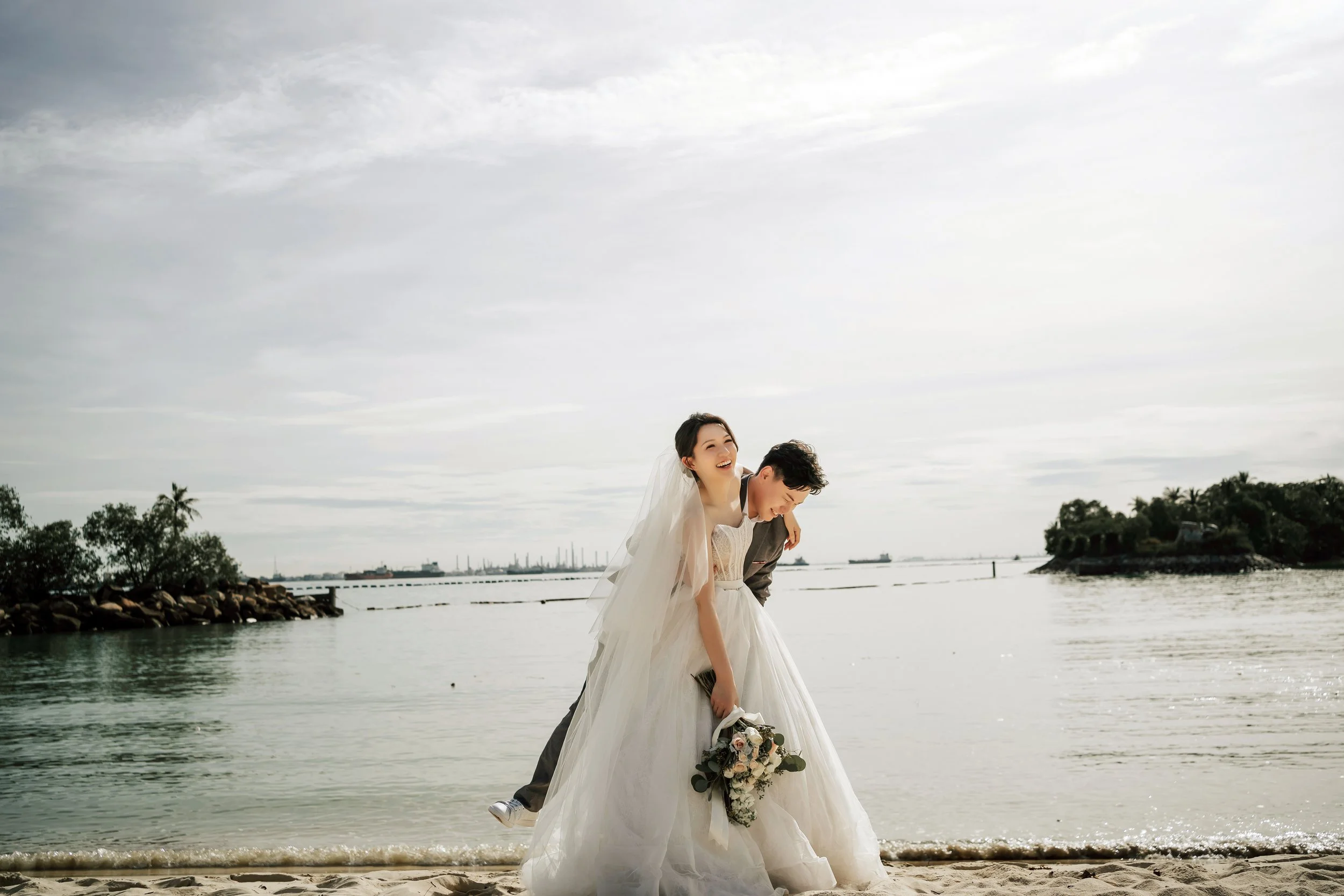 At Sentosa Beach in Singapore, renowned for its serene sea views and scenic beauty, a couple in full wedding attire creates an unforgettable tableau in their pre-wedding photoshoot. The groom, strong and loving, is captured mid-attempt at lifting his