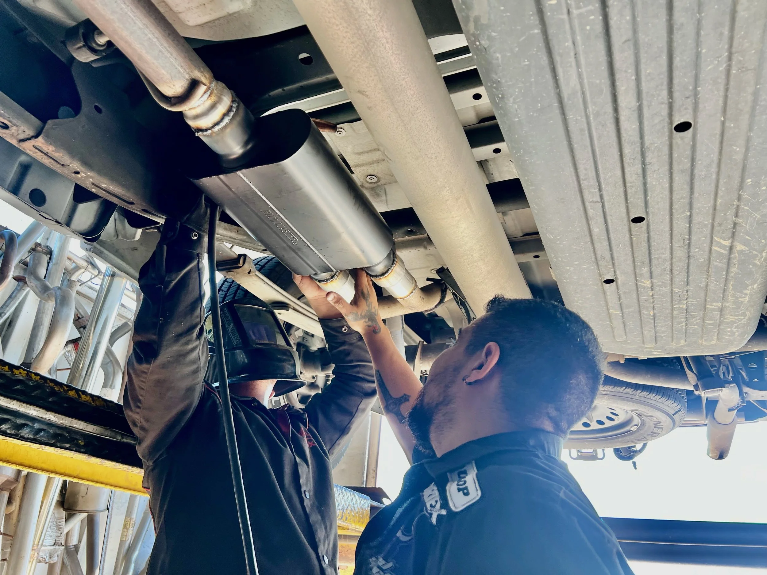 Two mechanics working underneath a vehicle, inspecting or repairing the exhaust system.