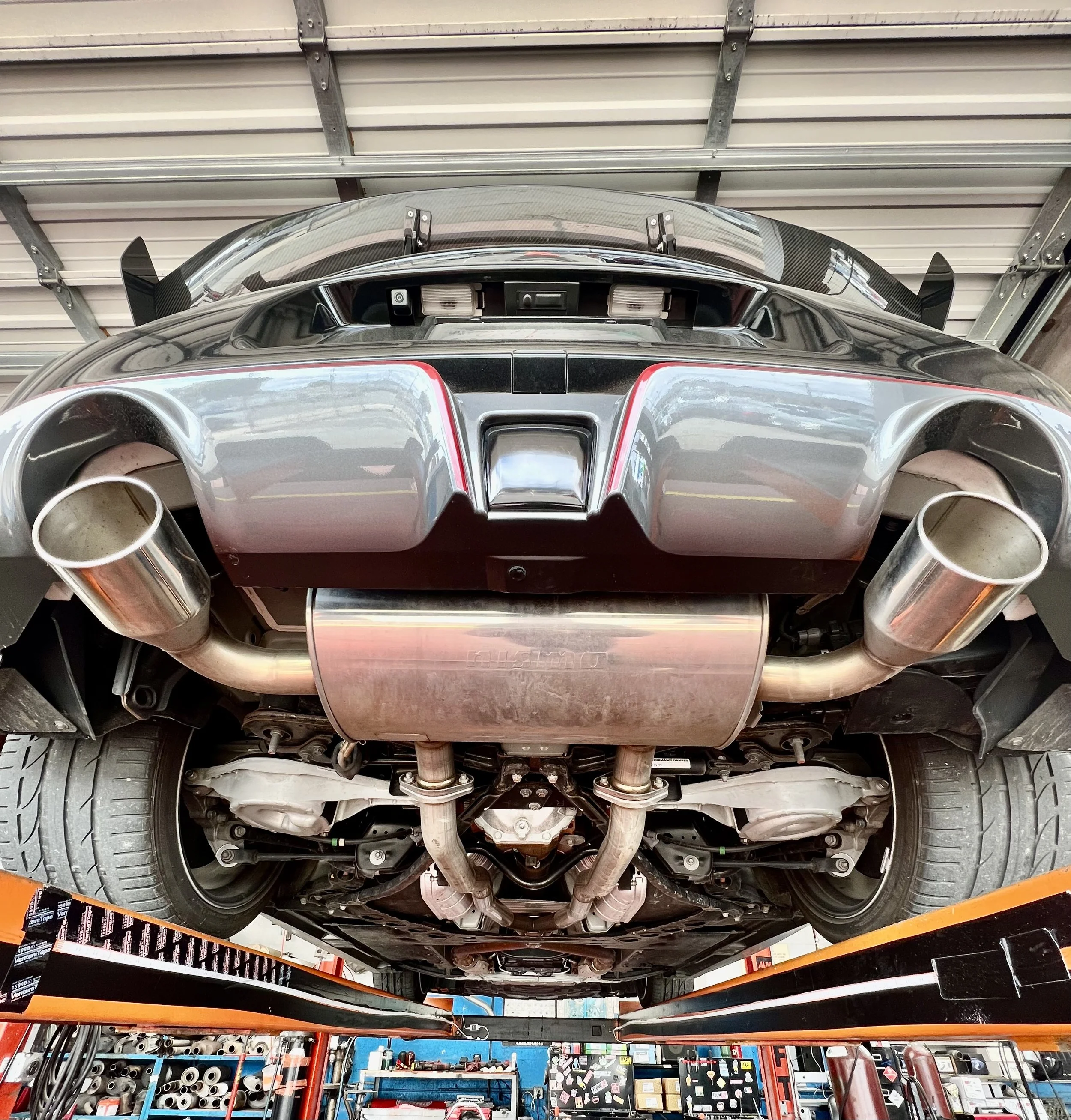 Underneath view of a sports car showing exhaust pipes, rear suspension, tires, and underside components in a garage or workshop