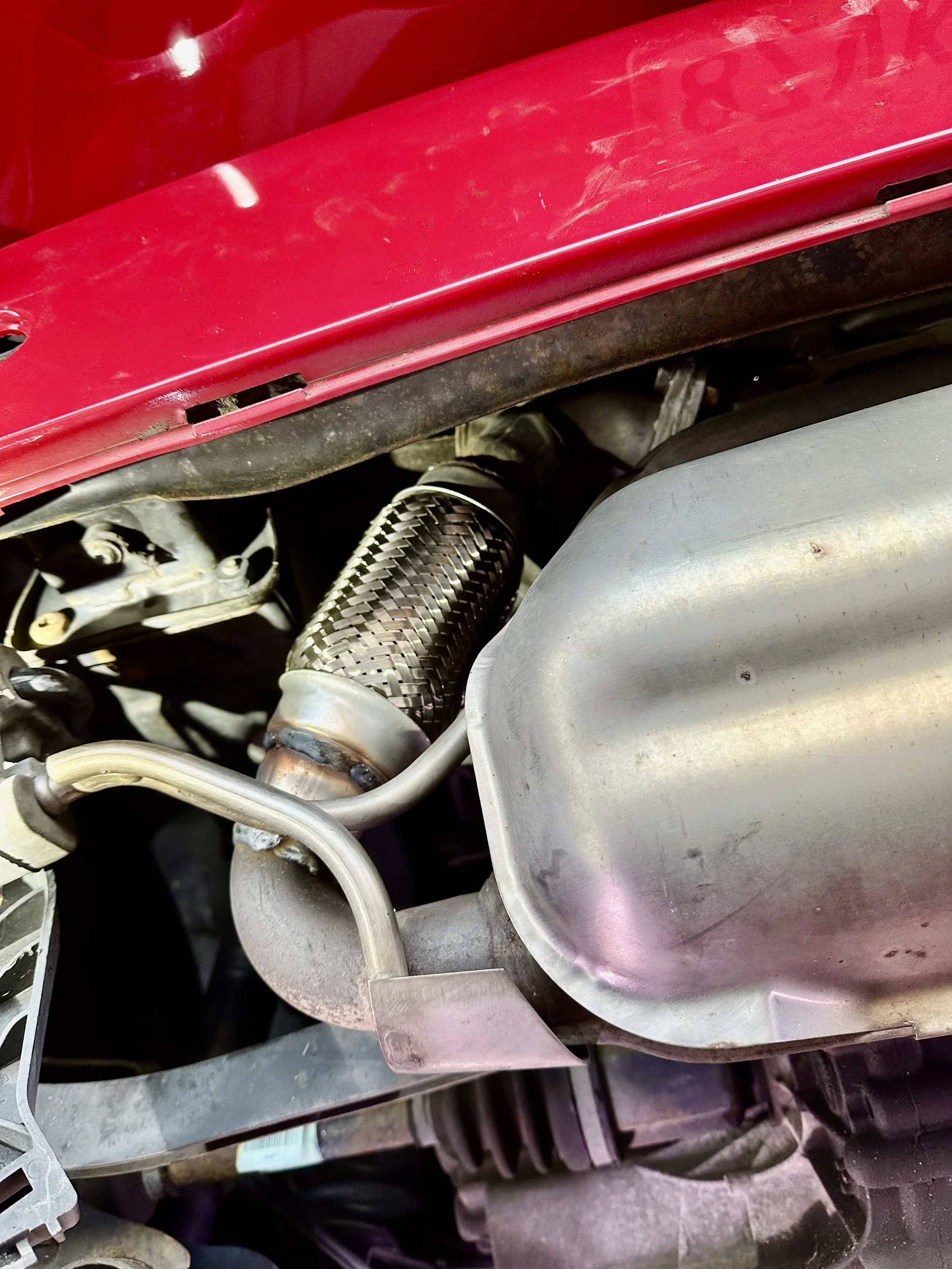 Close-up of a car engine showing a metallic exhaust pipe with a heat shield, an aluminum oil pan, and part of a red vehicle body.