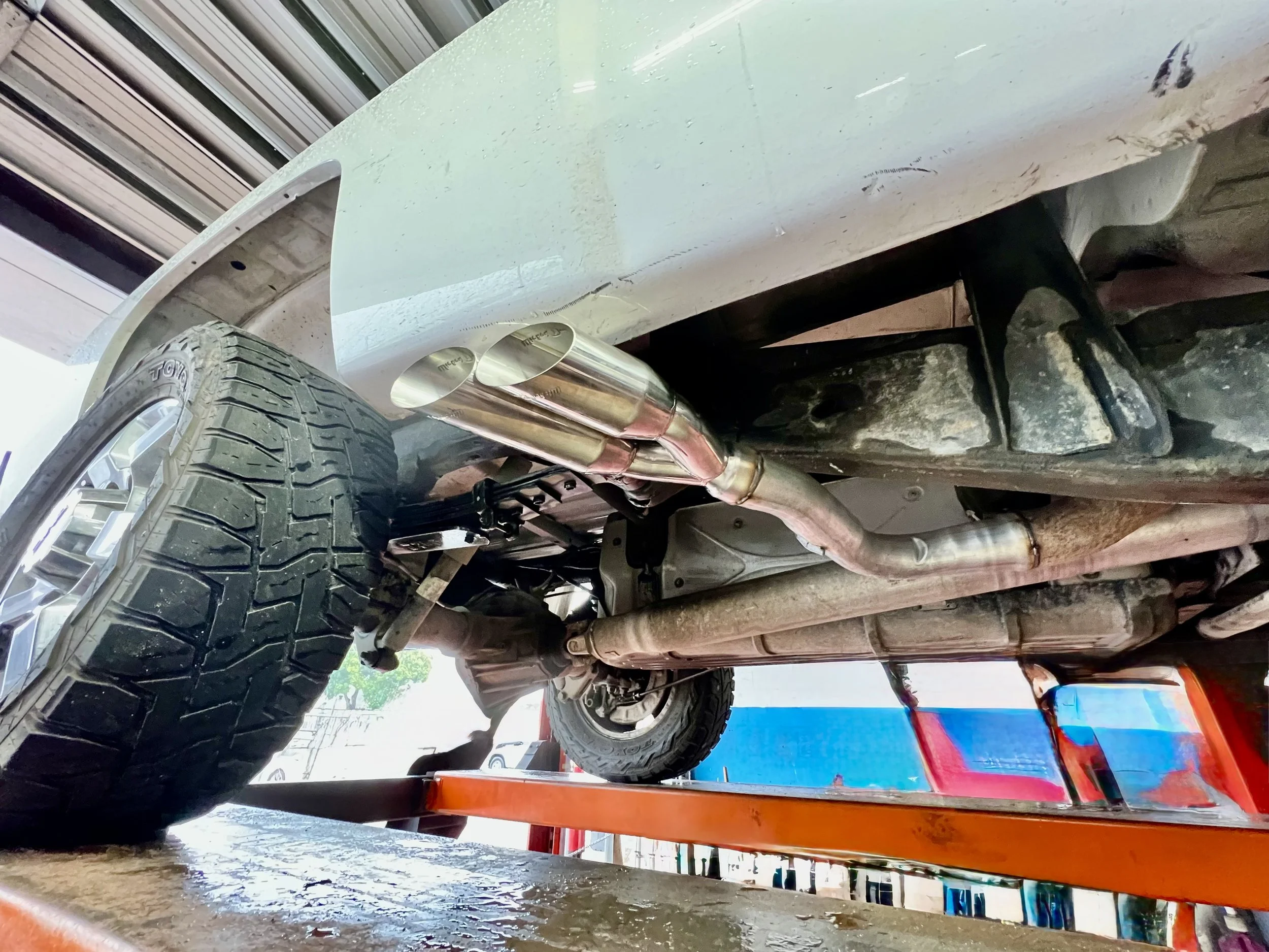 View of the underside of a vehicle showing exhaust pipes, suspension, and a spare tire, with the vehicle on a red lift in a garage or workshop.