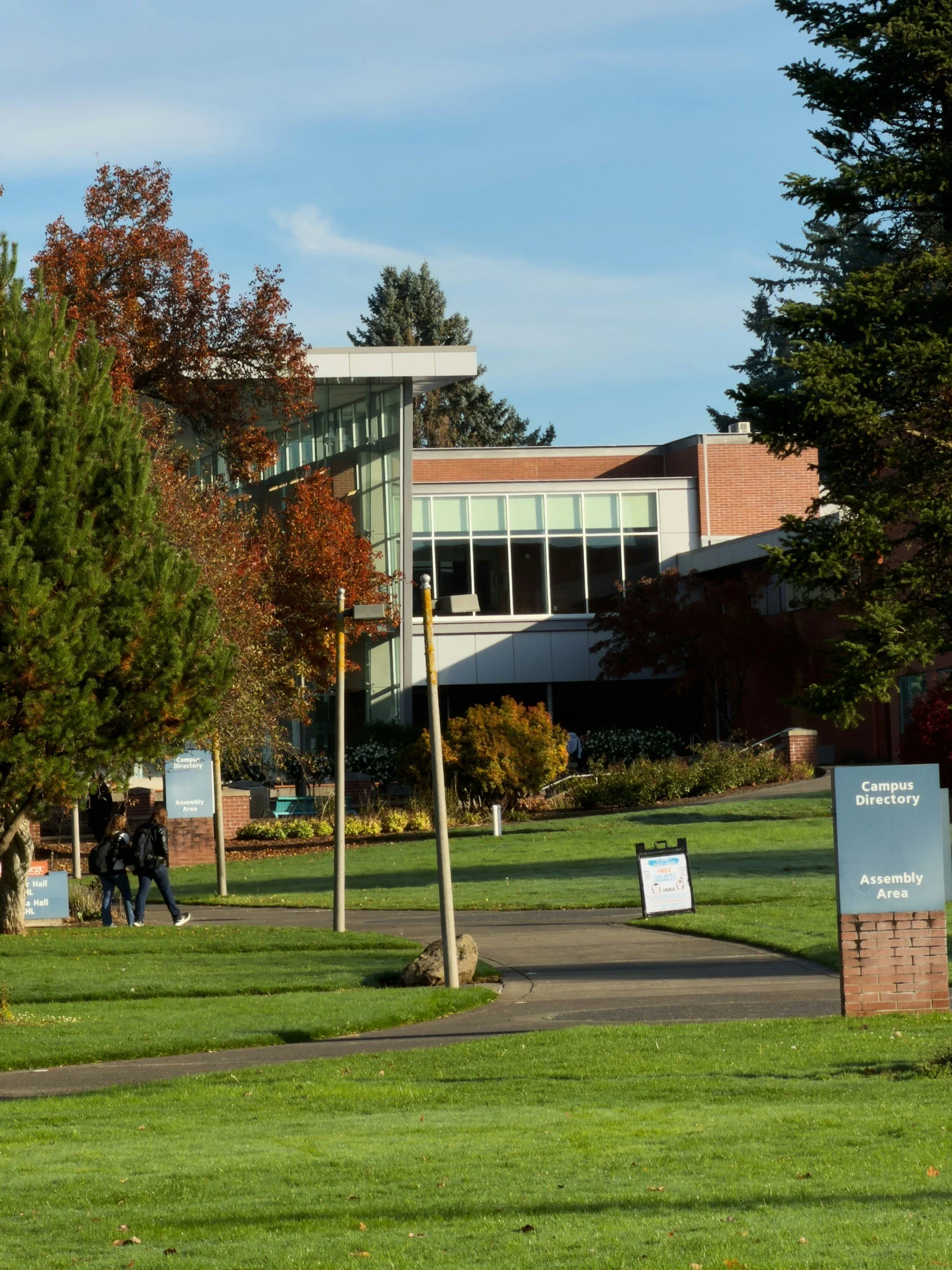 Campus with green lawns, trees with autumn leaves, campus signs, and a modern building with glass windows in the background.