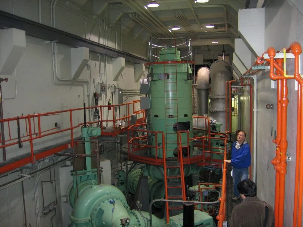 Industrial machinery inside a factory or power plant, with two men inspecting the equipment, surrounded by pipes and orange railings.