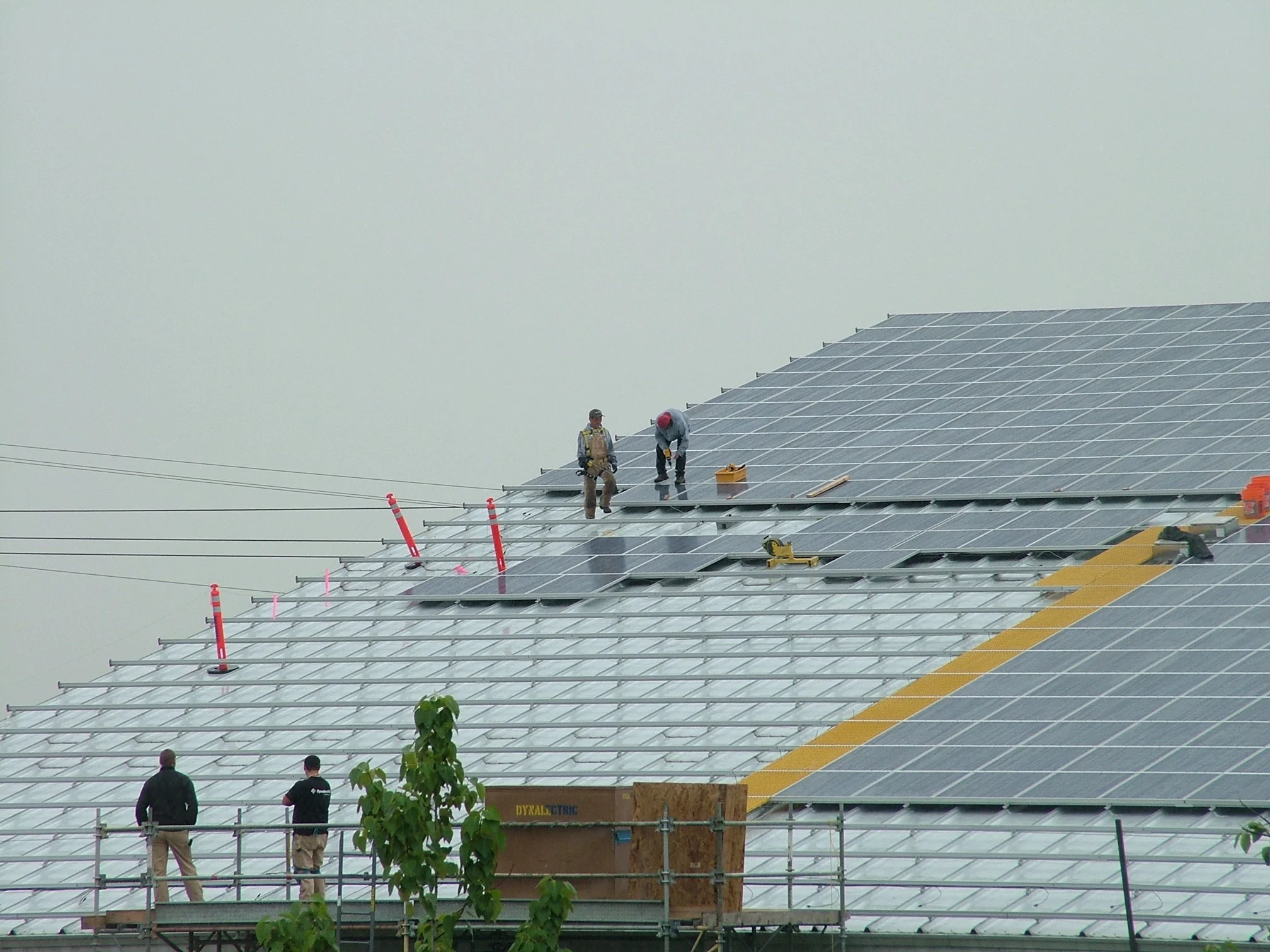 Workers installing solar panels on a large sloped roof, some standing on scaffolding, with foggy sky in the background.