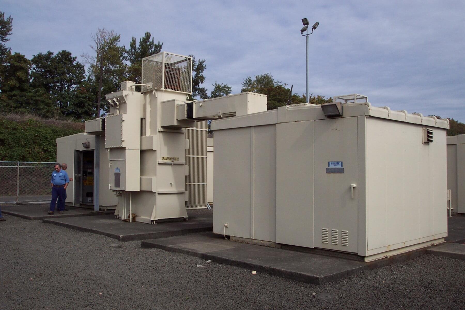 Outside of a secure utility area with large equipment and transformers, two people in security uniforms stand near the fencing and equipment.