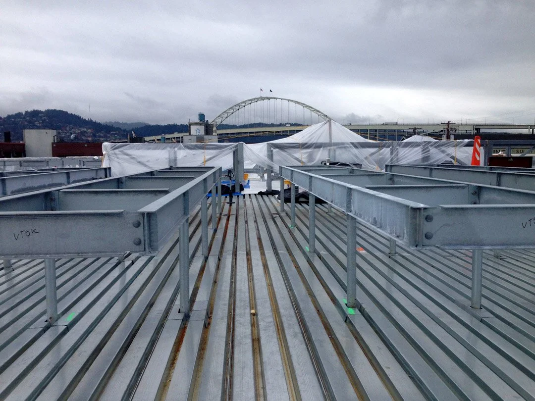 Rooftop construction with metal beams, covered with white plastic sheeting, overlooking a cityscape and a bridge in the distance, under cloudy sky.