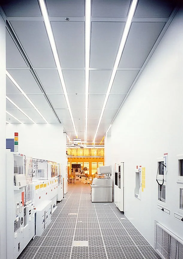 A bright, modern industrial or research facility with white walls, ceiling, and floor, equipped with various control panels, machines, and monitors along the sides, and illuminated by linear LED ceiling lights.