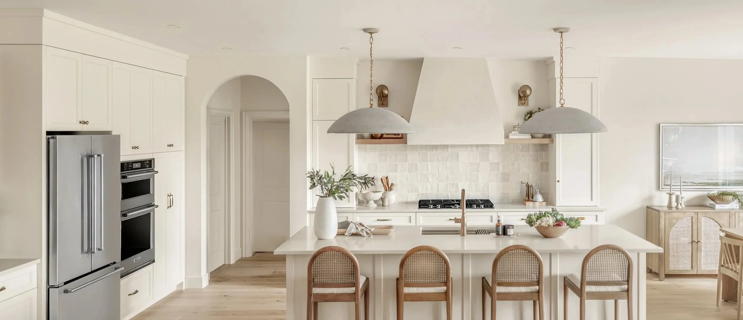 Bright kitchen with white cabinets, a large island with a white countertop, and three bar stools. Stainless steel refrigerator and oven, modern chandelier lights, and a decorative vase with greenery on the island. Light wood flooring and minimalist decor.