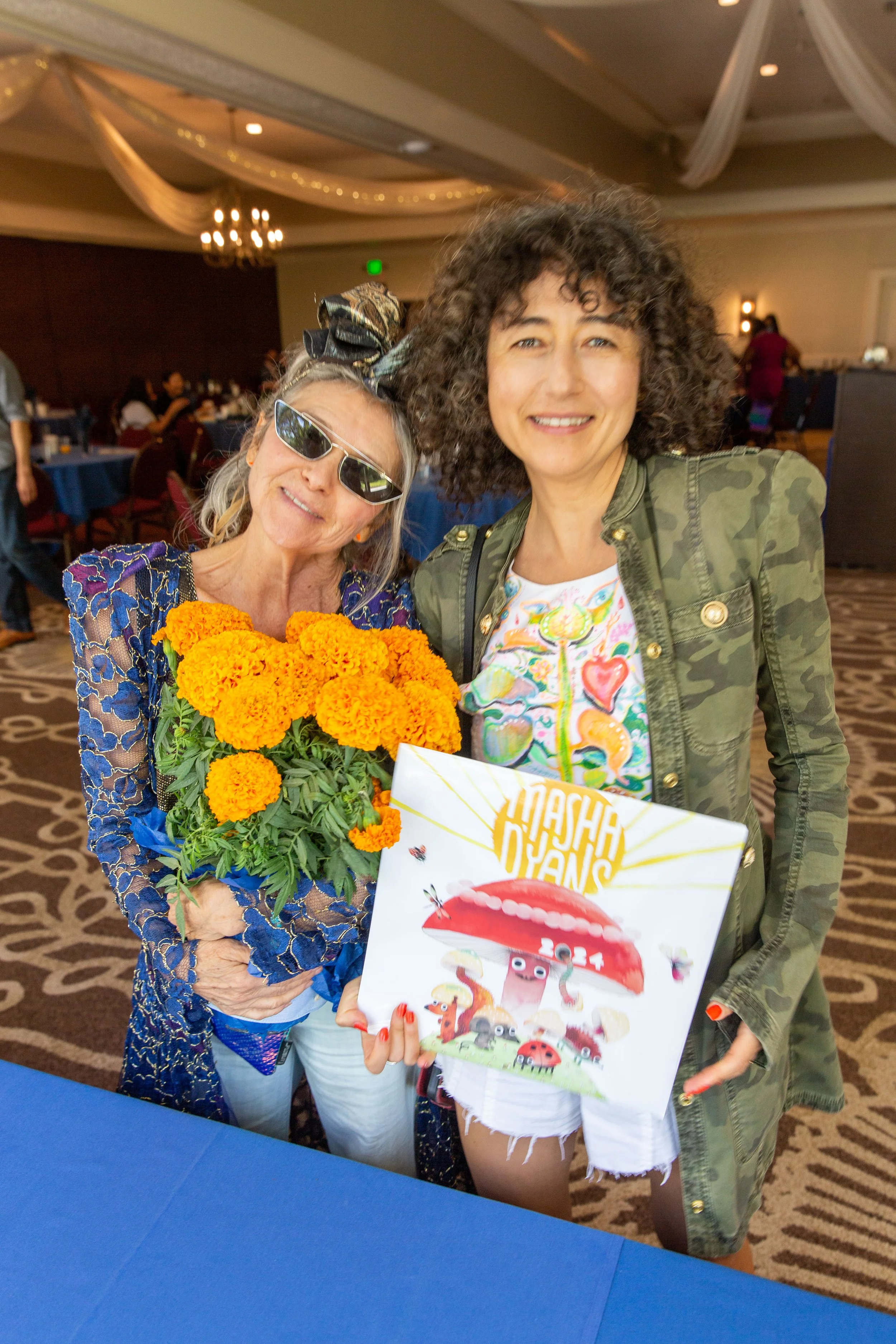 R to L: Award-winning author/illustrator Masha D'yans holds up her 2024 calendar beside her mother who is holding a marigold floral centerpiece. (Josh Steichmann)