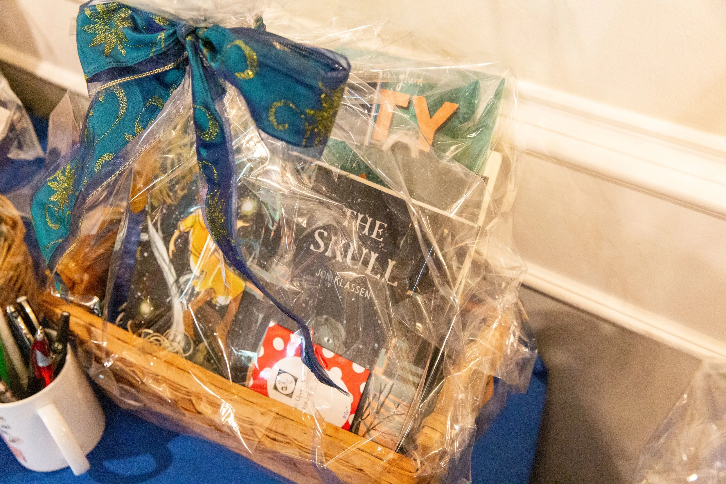 A cup of pens nestles next to a silent auction basket including picture books by Southern California creators Rebecca Jordan-Glum (Kitty), Kirsten W. Larson (The Fire of Stars), and Jon Klassen (The Skull). These titles were all recent CLCSC Book Clu