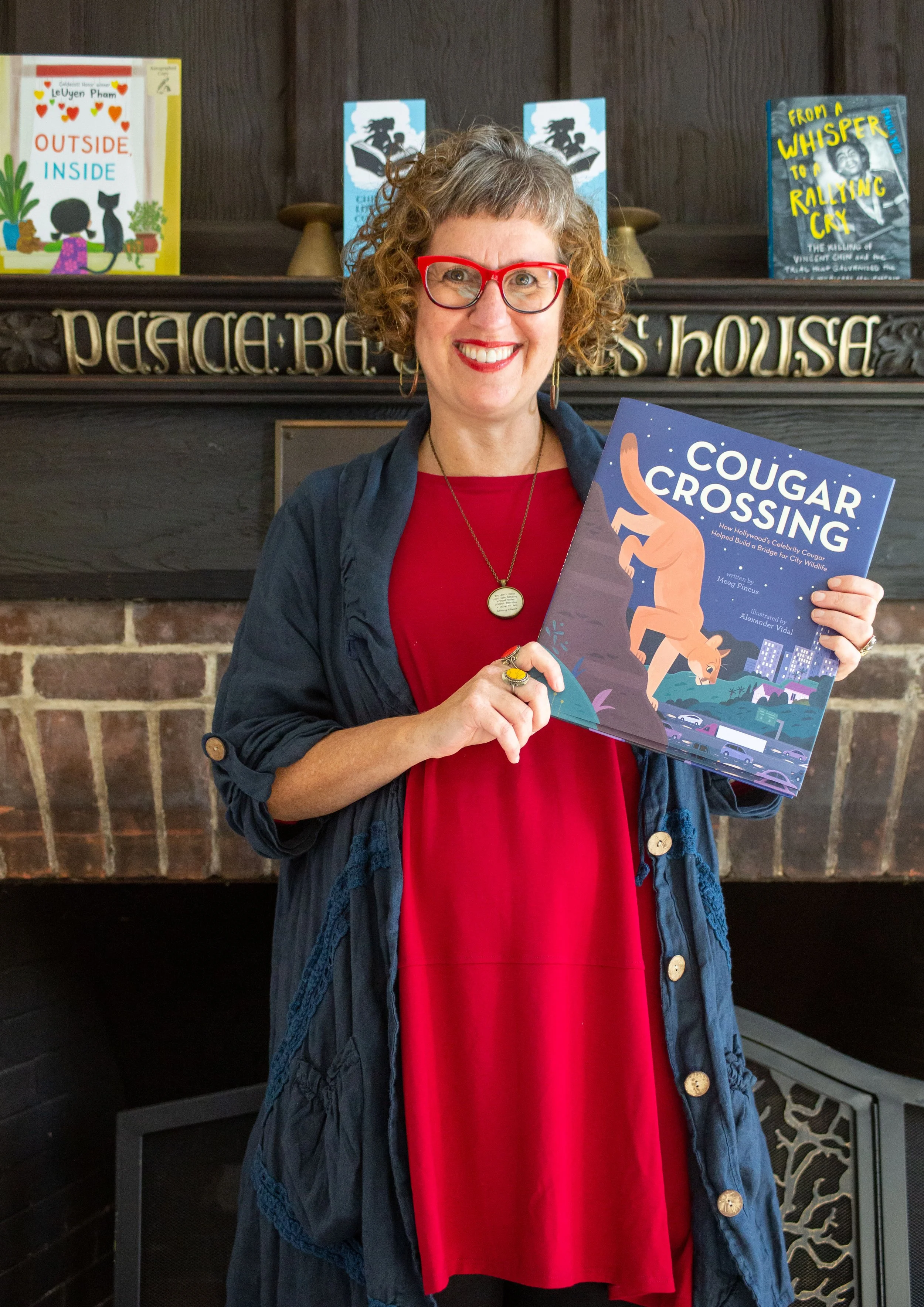Meeg Pincus stands before a fireplace holding a copy of her book Cougar Crossing, illustrated by Alexander Vidal. Photographed by Josh Steichmann on November 12, 2022 at Throop Church in Pasadena, CA.