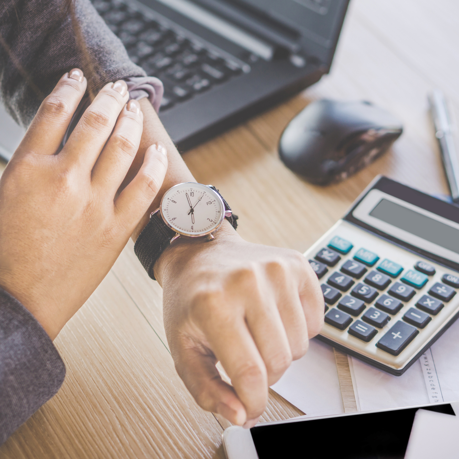Person checking wristwatch next to laptop, calculator, and smartphone on desk.