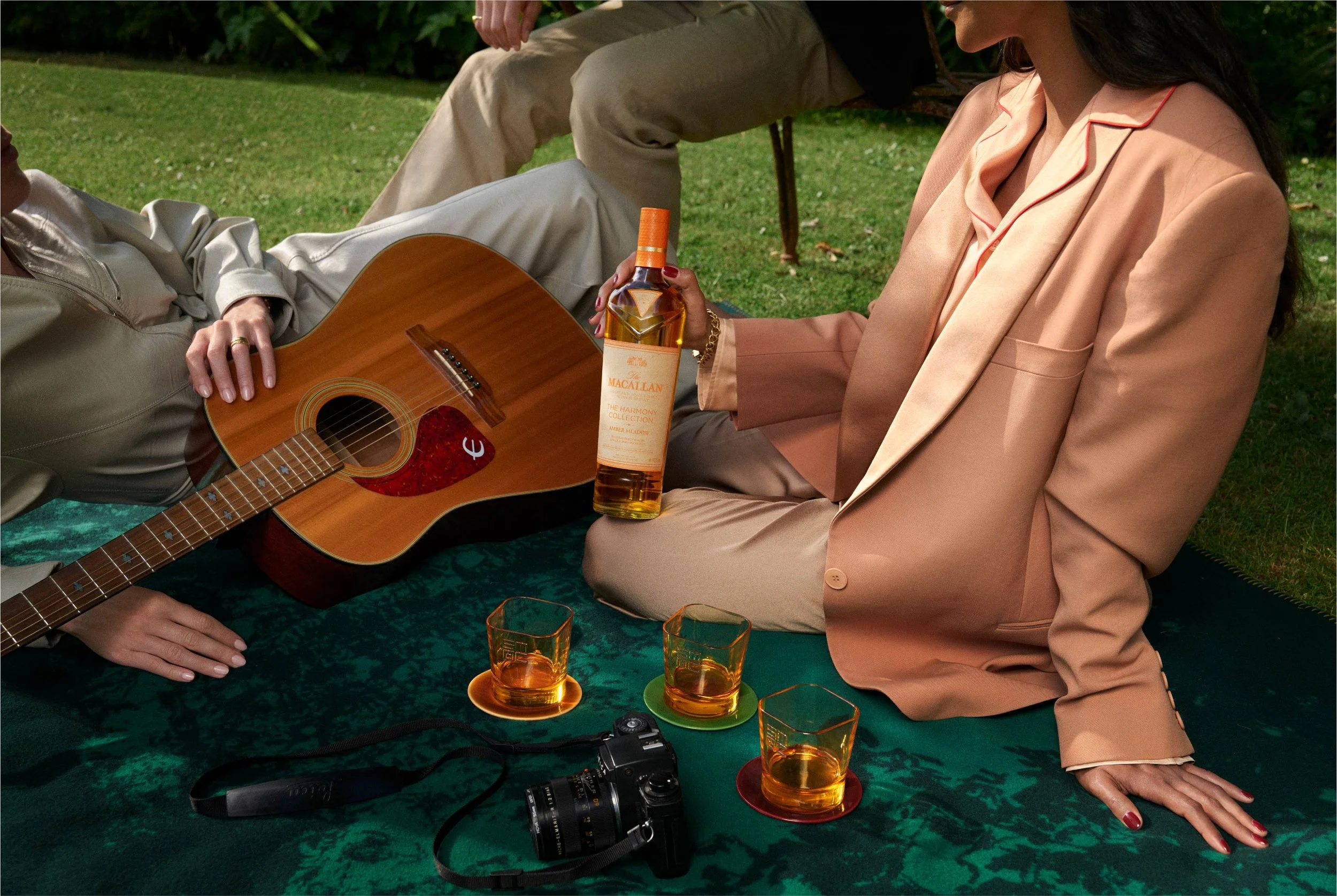 People sitting on a green surface outdoors with drinks, a guitar, and a camera, engaged in a social gathering.