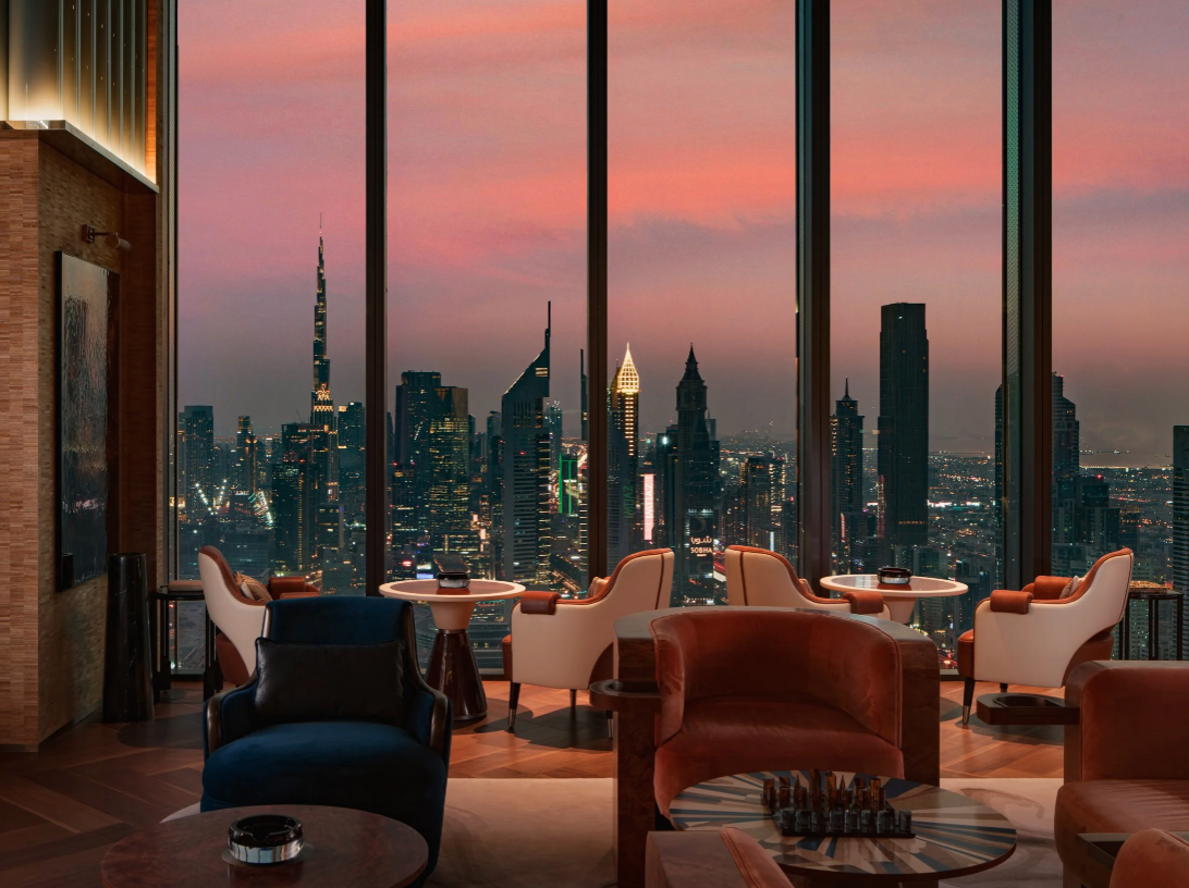 Luxury lounge with large floor-to-ceiling windows overlooking a city skyline at dusk, featuring illuminated skyscrapers and a pink and purple sky, furnished with modern chairs and tables.