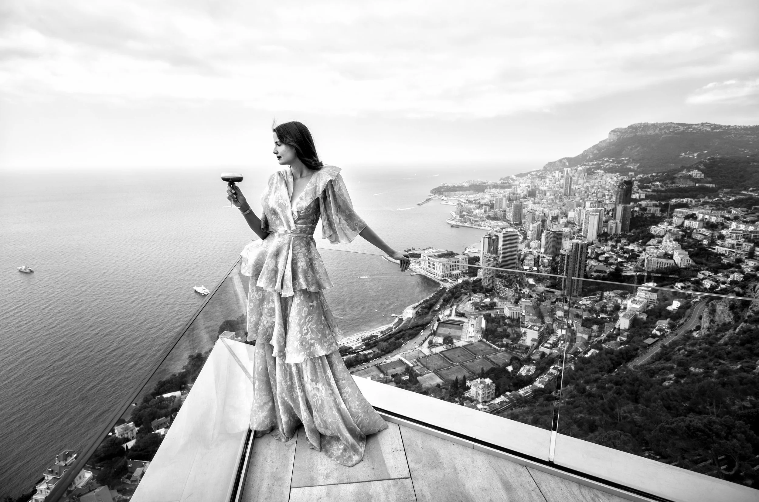 A woman in a patterned gown standing on a glass ledge overlooking a city and body of water, holding a cocktail glass.