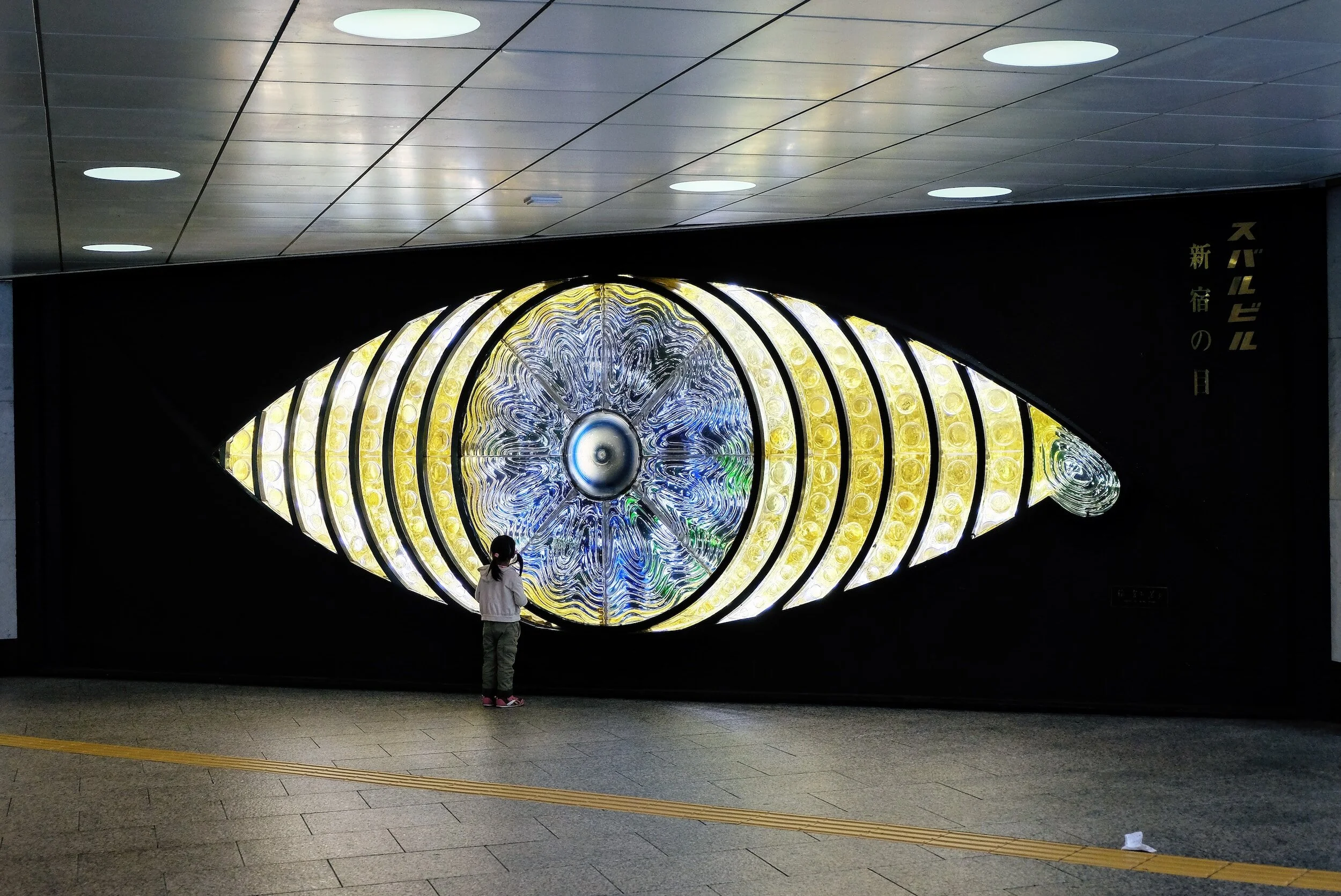 A child standing in front of a large eye-shaped artwork with illuminated circular patterns inside, located in an indoor public space.