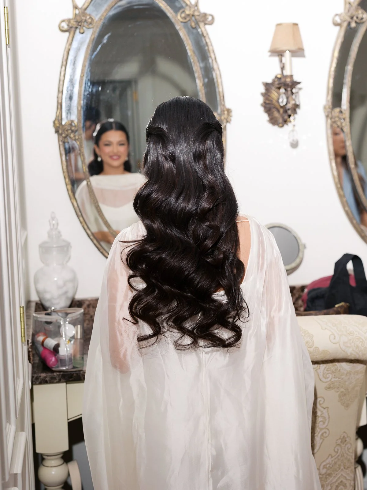 A Latina bride with full glam hair and makeup done wearing a sheer robe, looks at herself in the mirror before putting on her wedding dress.