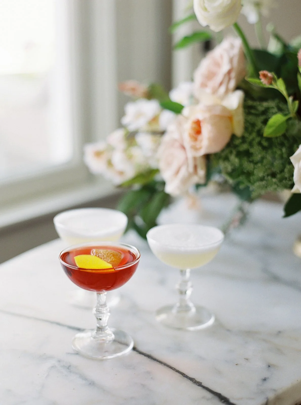  Beautiful hand crafted cocktails sit on a marble countertop near a window and flower arrangement. 