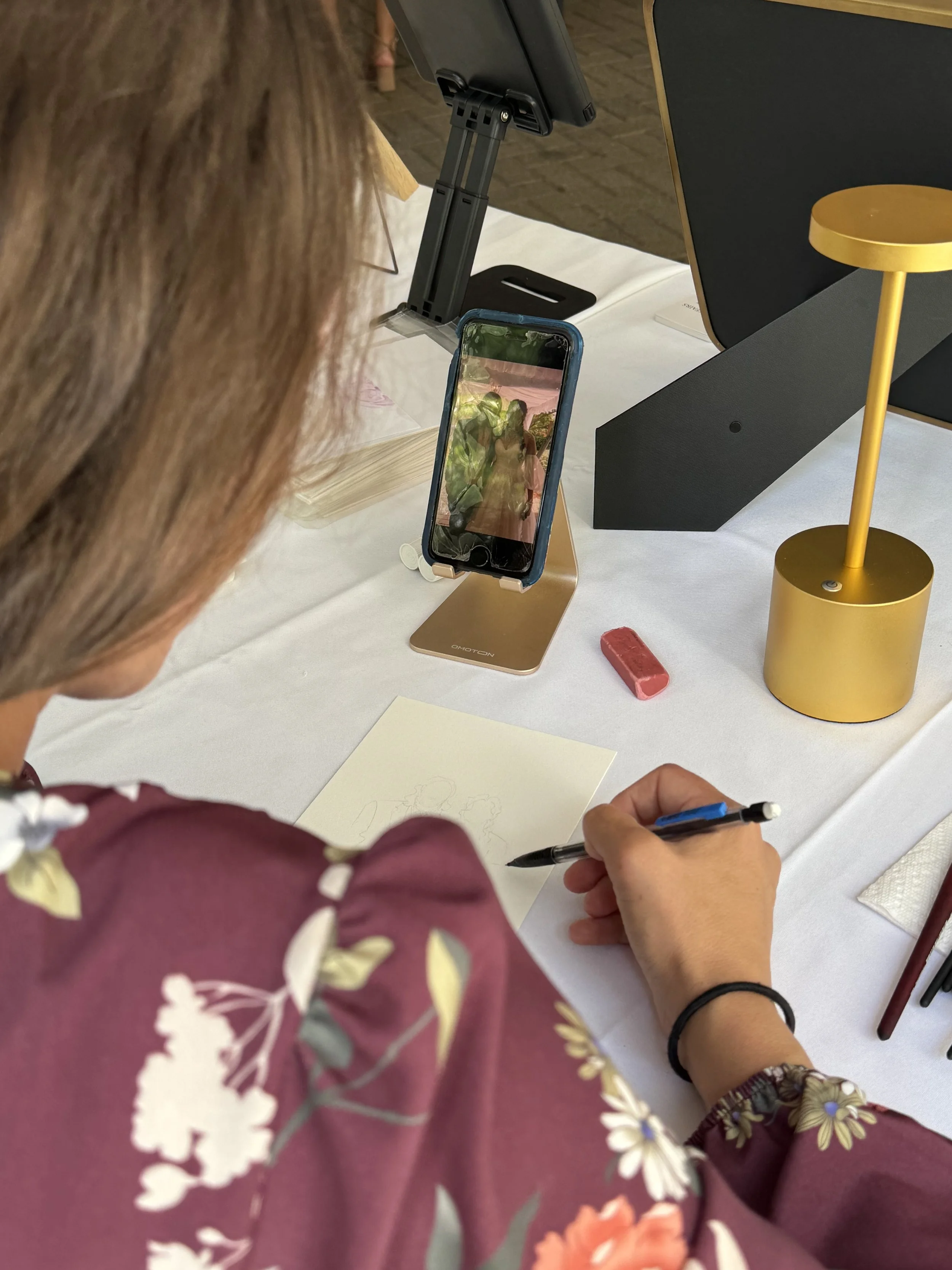 A wedding guest illustrator begins to sketch a young couple in real time onto a piece of watercolor paper at a table with a small lamp.