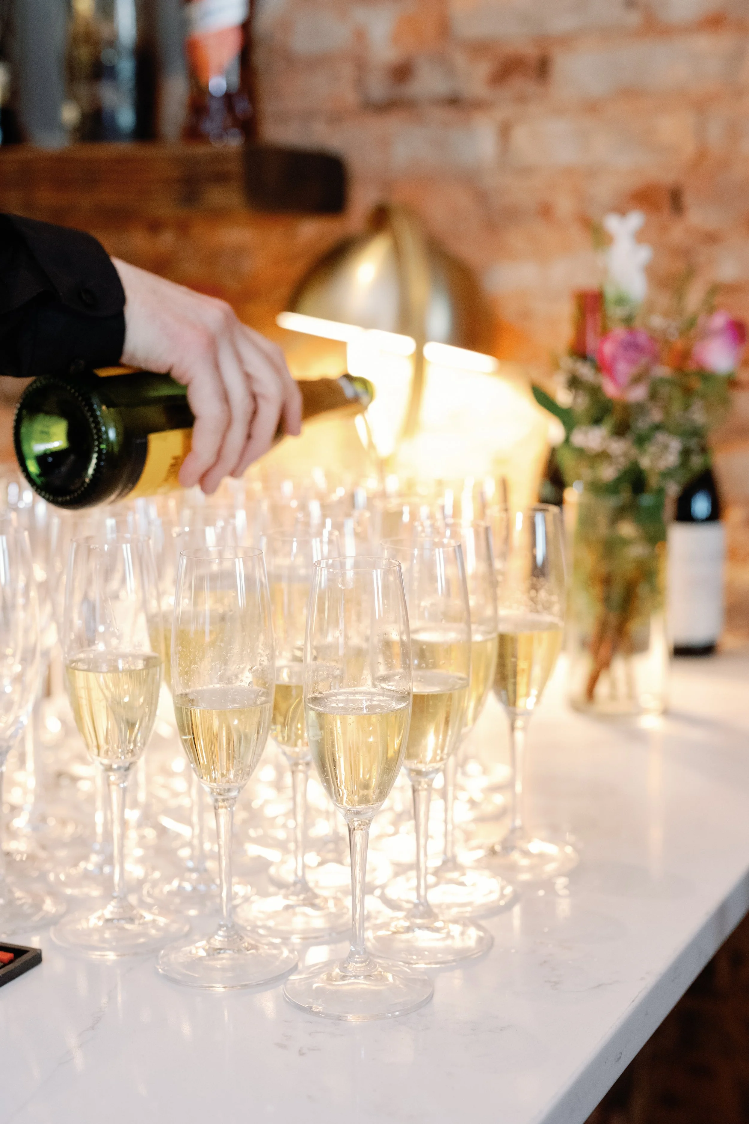 A server at a high end catered event pours champagne into tall, sparkling flutes.
