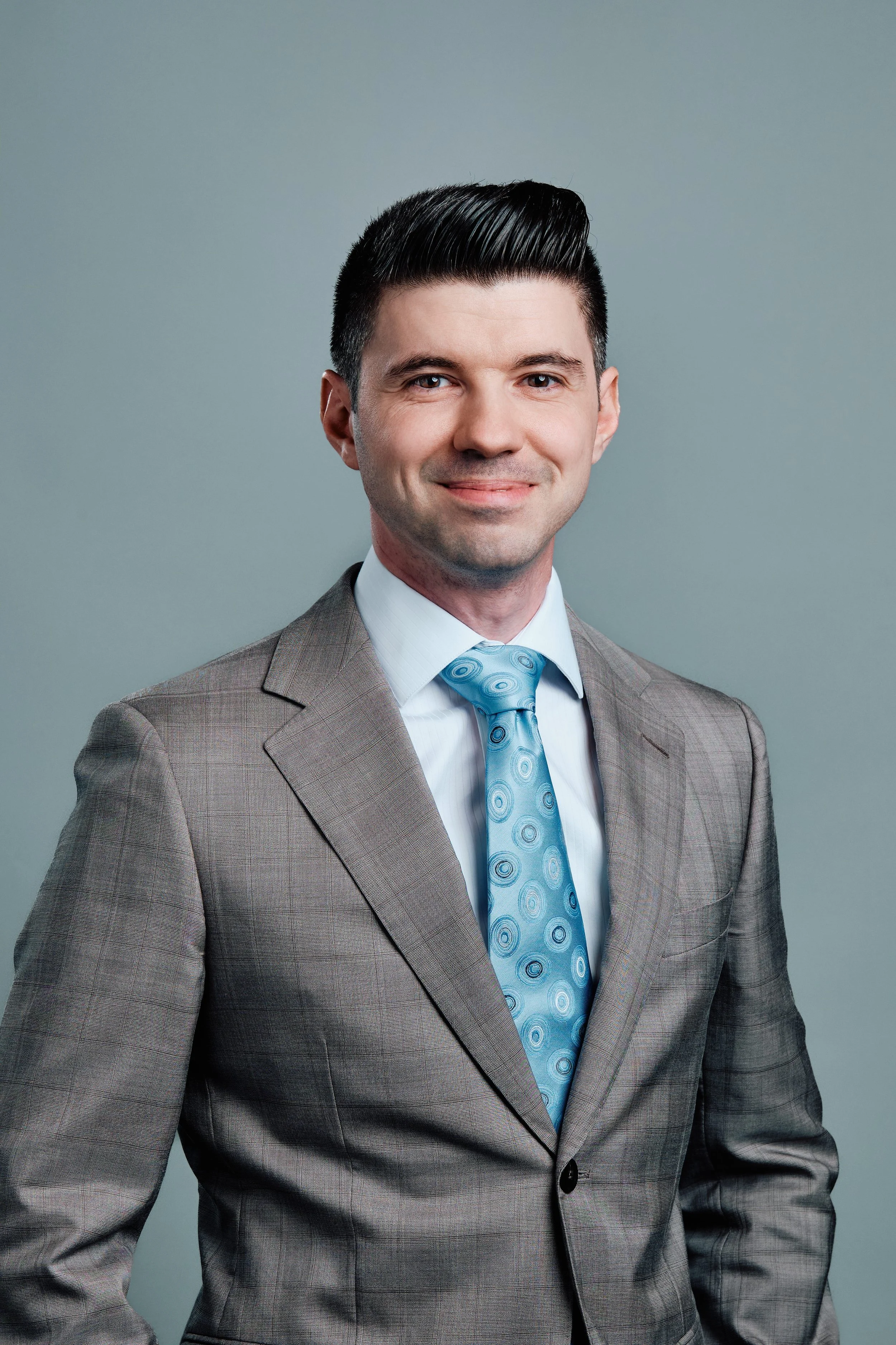 A portrait of a man in a gray suit with a blue patterned tie, standing against a gray background, smiling at the camera.