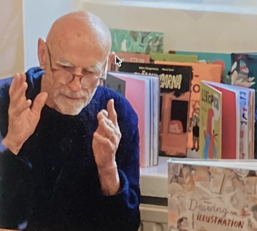 An elderly man with glasses and a white beard, wearing a dark sweater, is gesturing with his hands while sitting in front of a table filled with colorful books.