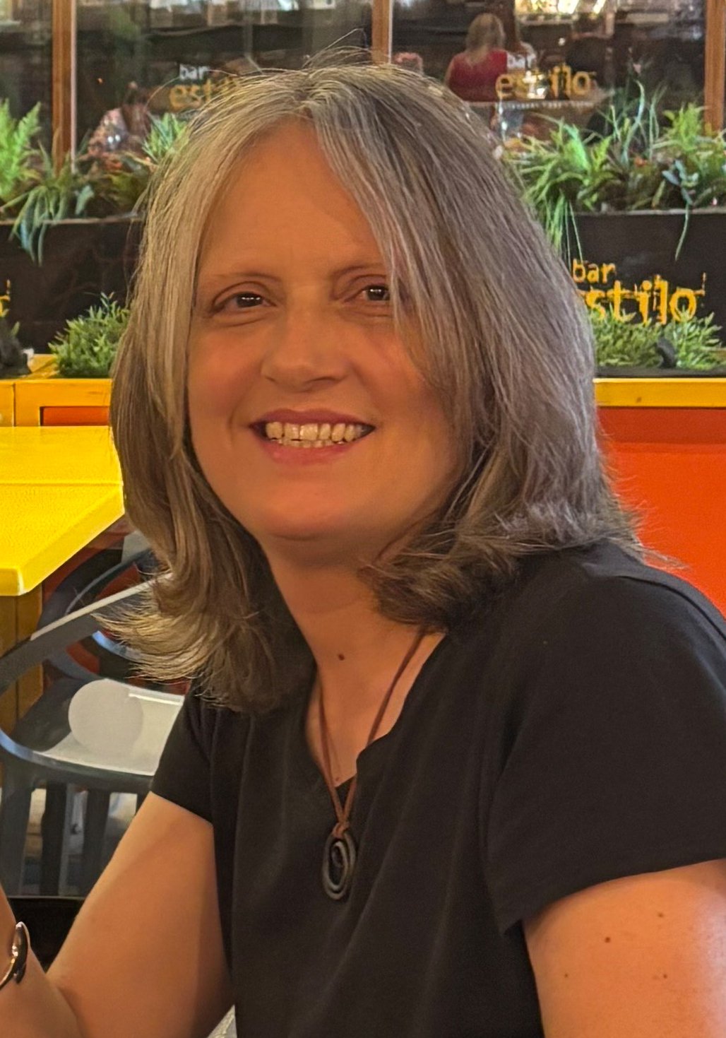 A woman with shoulder-length gray hair smiling at a restaurant with colorful tables and plants in the background.