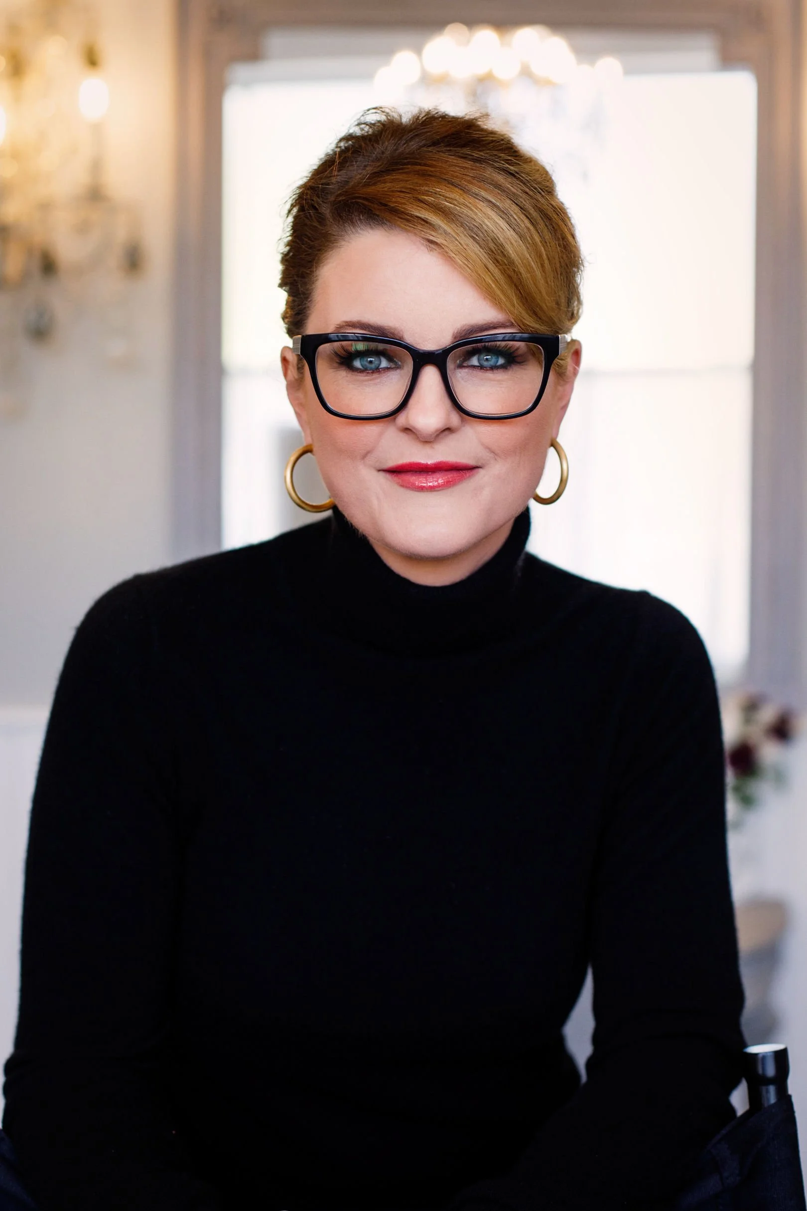 A woman with red hair, wearing glasses, gold hoop earrings, and a black turtleneck, smiling indoors with a chandelier and window in the background.