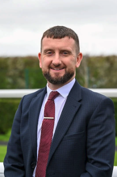 Man with short dark hair and beard wearing a navy suit, white shirt, and maroon tie standing outdoors with a blurred green landscape in the background.
