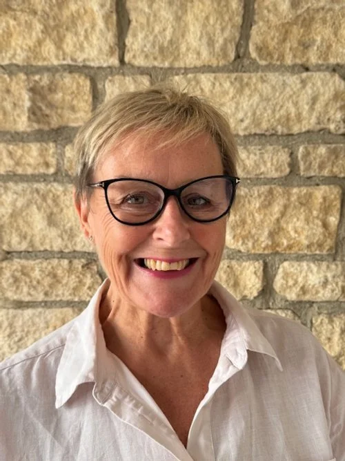 Close-up of a smiling woman with short gray hair, wearing glasses and a white collared shirt, standing in front of a textured brick wall.