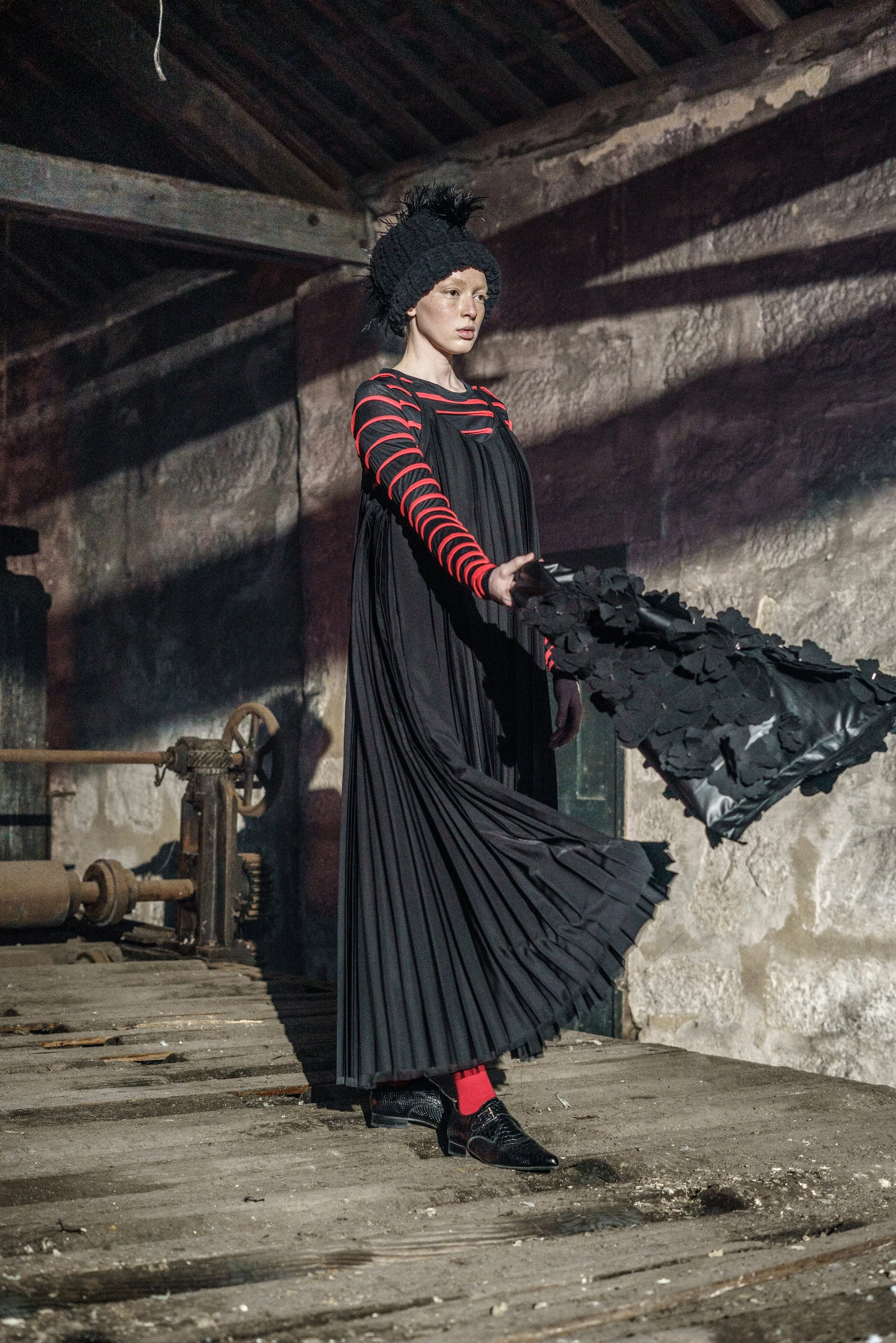 A woman in a black and red striped shirt, black pleated dress, and black hat with feathers, holding a black umbrella with ruffled fabric, standing in an industrial setting with a textured stone wall and wooden floor.