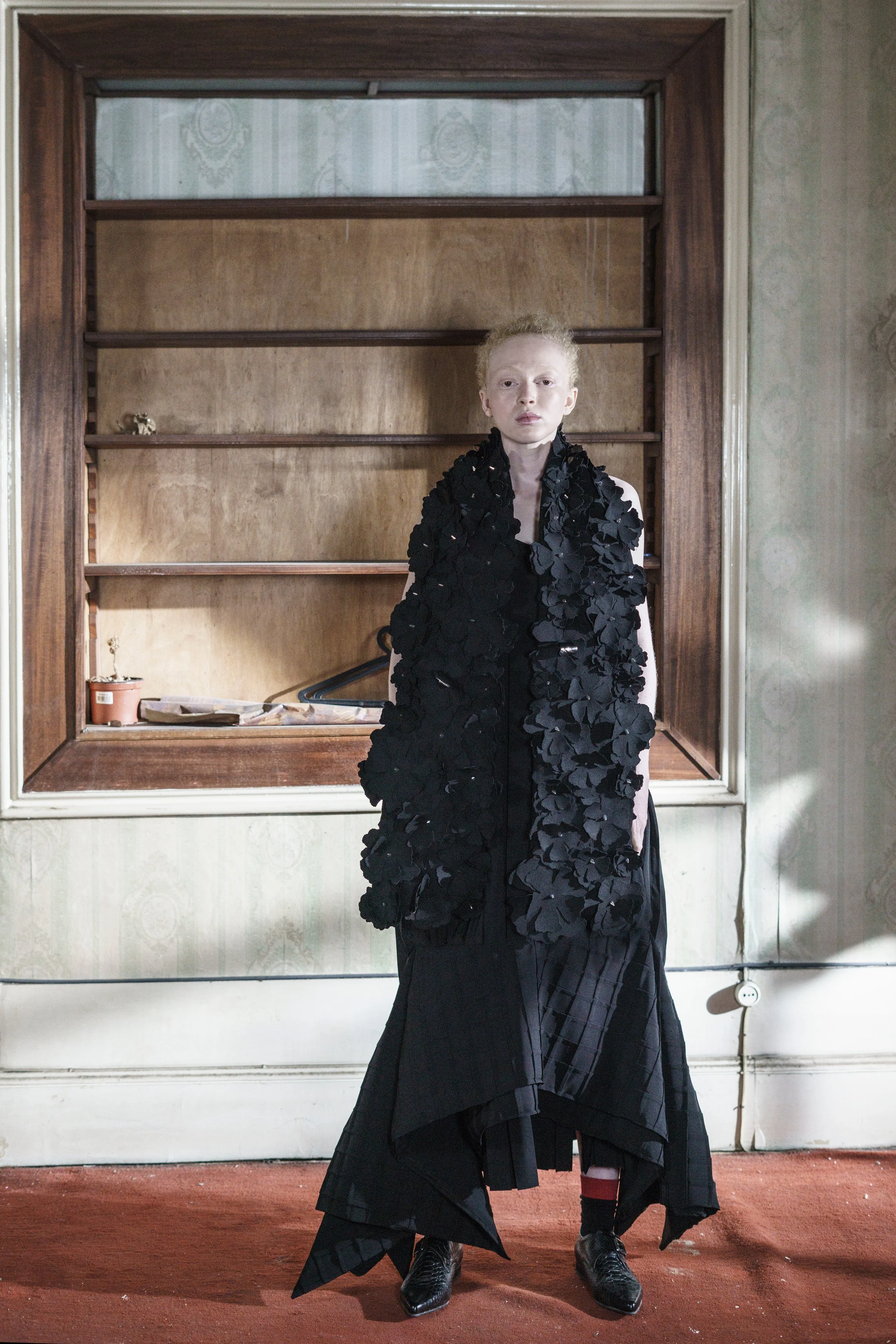 A person with pale skin and short, curly blond hair stands in front of an empty wooden bookshelf, wearing a black textured outfit and black shoes, in a room with greenish patterned wallpaper and a red carpet.