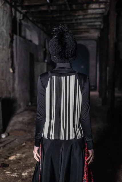 Person walking through a dark, abandoned industrial space wearing a striped vest, black pants, and a fuzzy black hat.