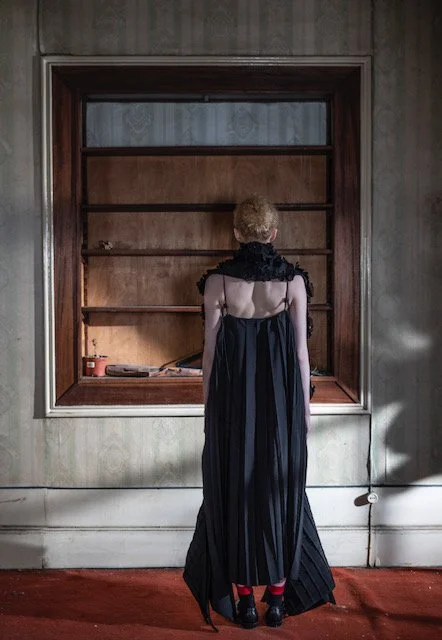 A person with short, curly hair wearing a black dress with sheer shoulder details stands with their back to the camera, facing an empty wooden bookshelf in an empty room with wood-paneled walls.